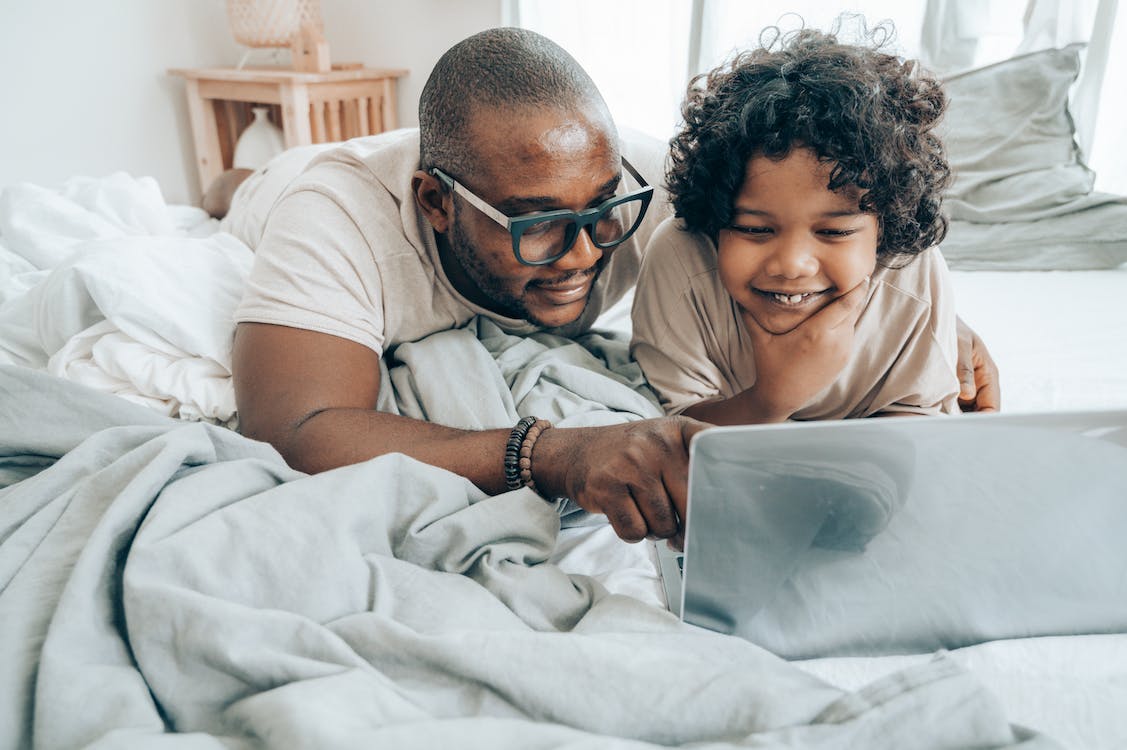 A black man and his son are looking at their computer while lying in bed together on a lazy Sunday, presumably enjoying transmedia storytelling experiences