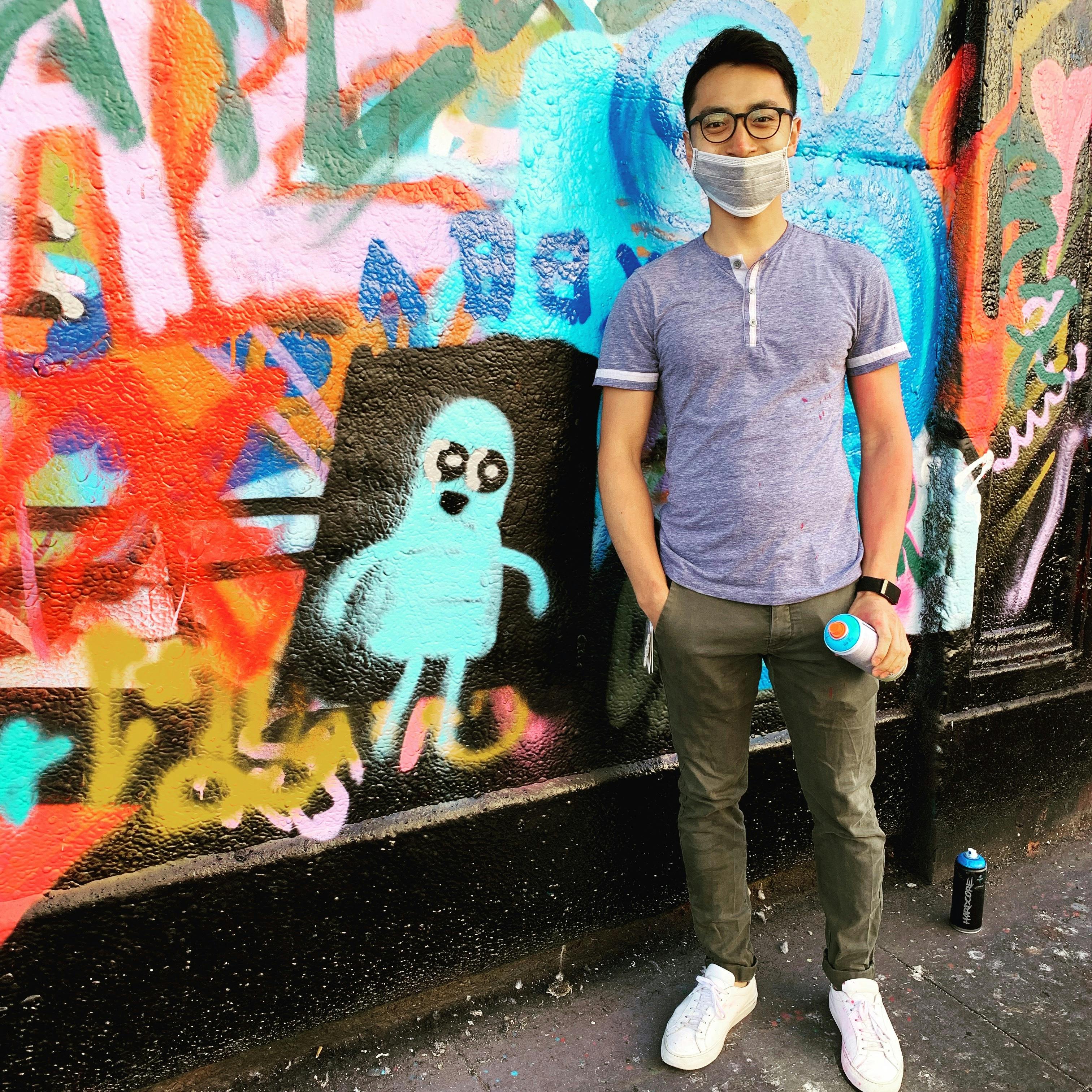 Alex, in a mask, is standing next to a character he drew with spray paint on a wall