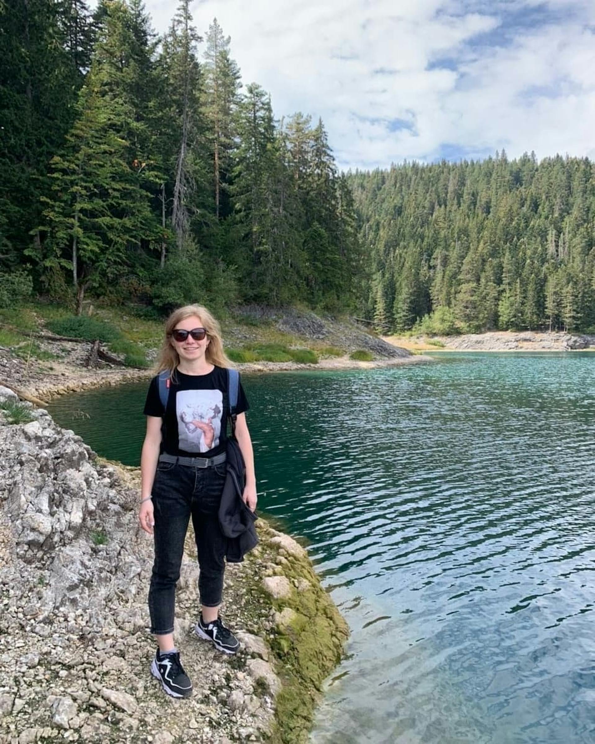 The woman is standing be the lake and a forest