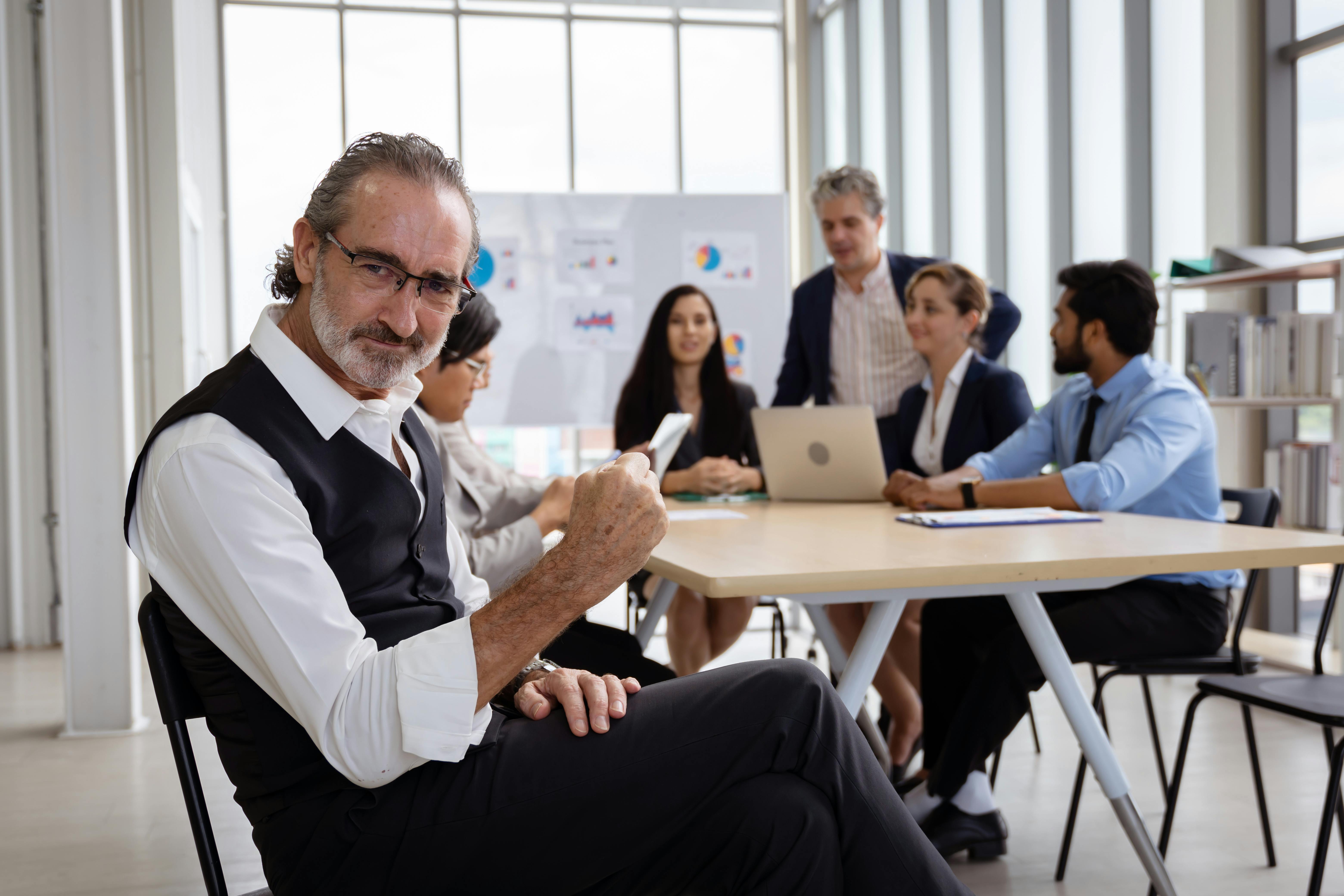 Ein Geschäftsmann zeigt sich selbstbewusst, während im Hintergrund sein Team in einem modernen Besprechungsraum an innovativen Geschäftsmodellen arbeitet.