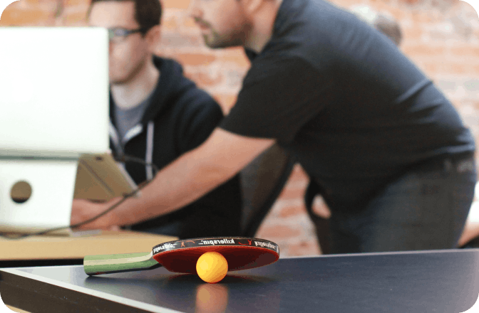 Ionic founders Max Lynch and Ben Sperry looking at computers with an in-focus ping-pong table with a dormant ball and paddle in the foreground