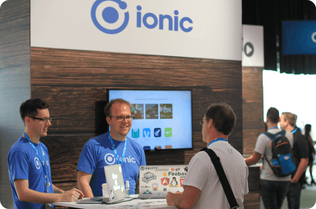 Ben Sperry and Perry Govier standing at a desk at an Ionic table behind Mac laptops, talking to a man