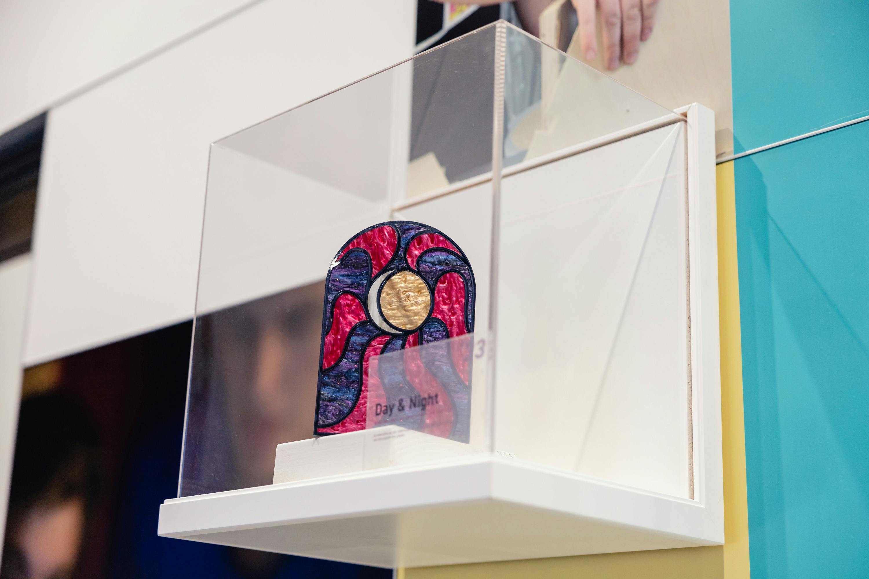 A colorful artistic object sits in a plastic exhibit box on a shelf