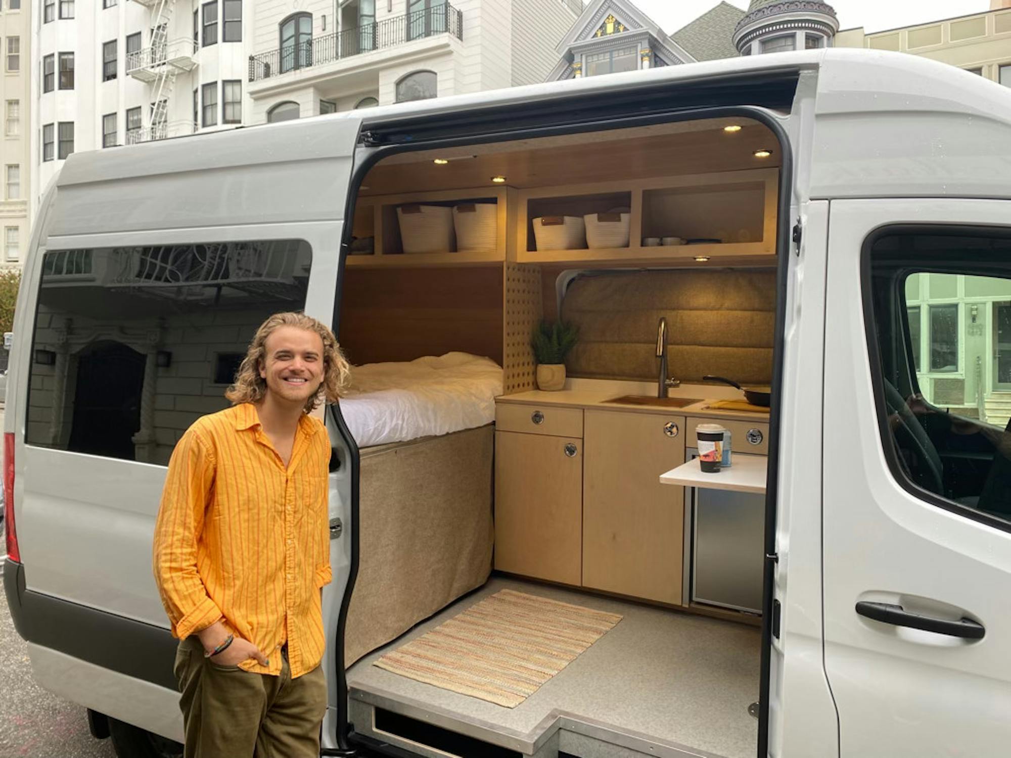 A young man in a loose yellow shirt stands smiling next to a van with an open door, revealing a furnished space inside with bed, sink, and small table.
