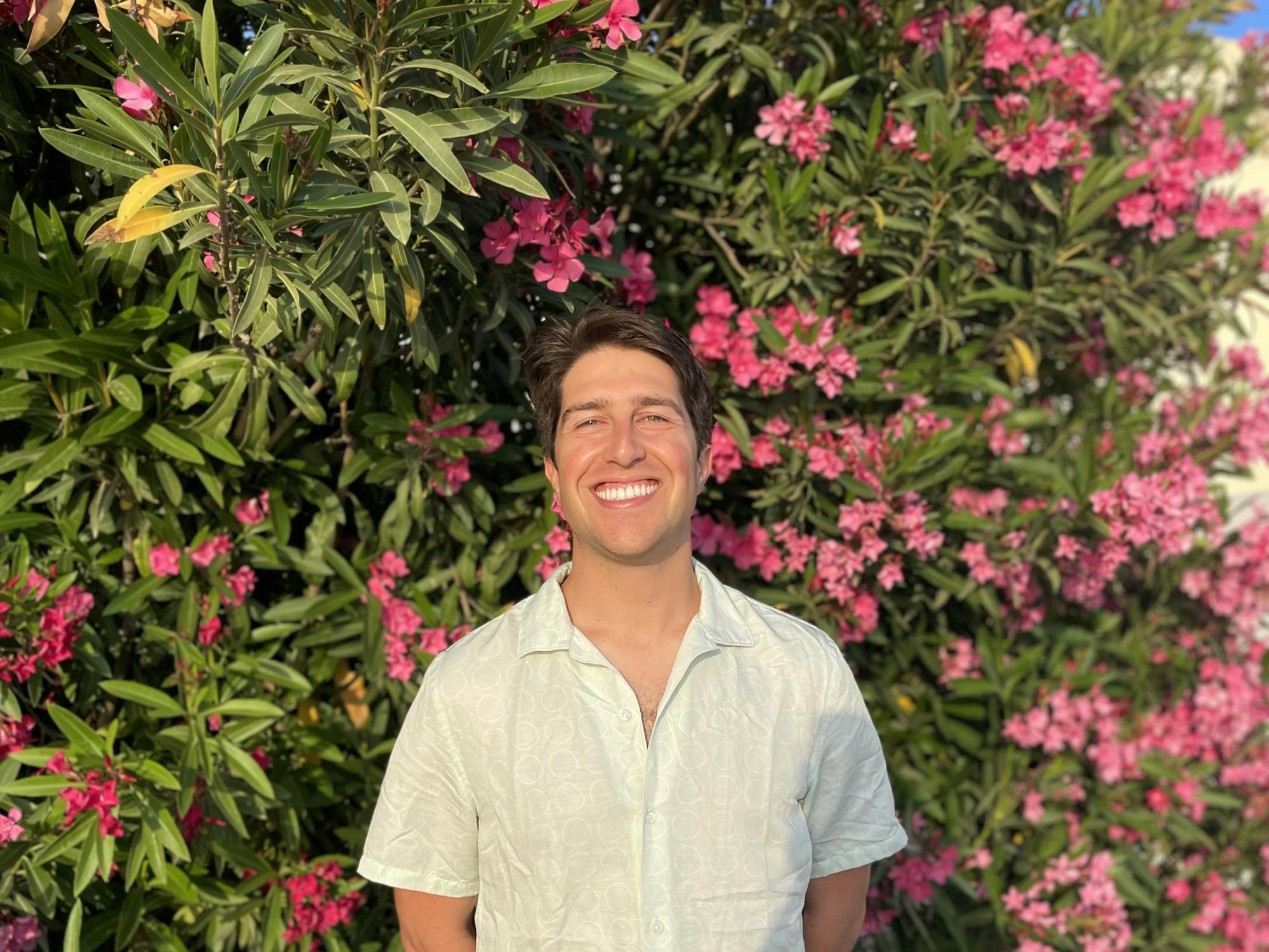 A man in a white button down short sleeved shirt smiles in front of pink flowers.