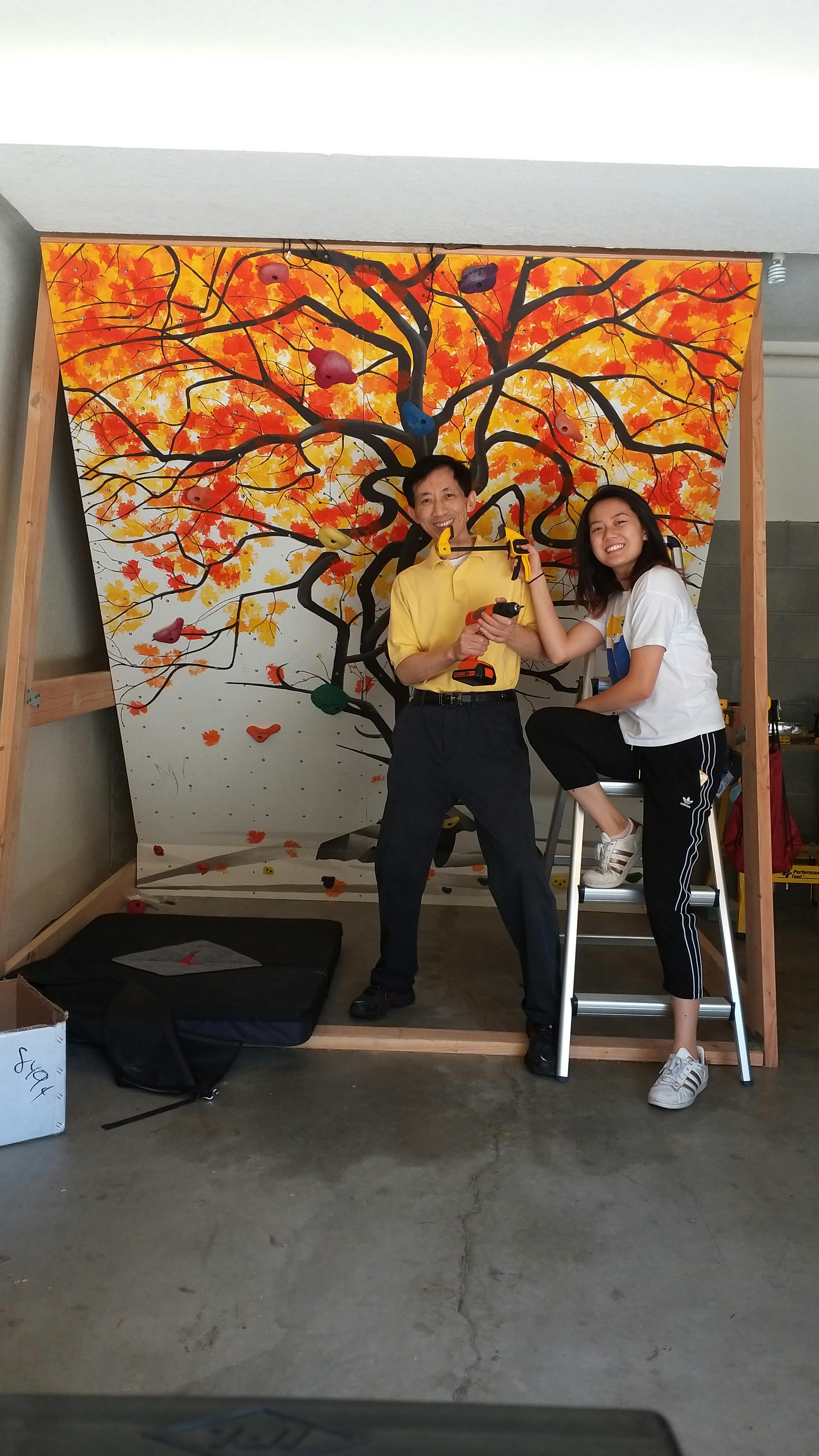 A young woman holding a clamp poses over an older man holding a drill in front of a person-sized canvas painted with an orange-and-yellow-leafed tree.