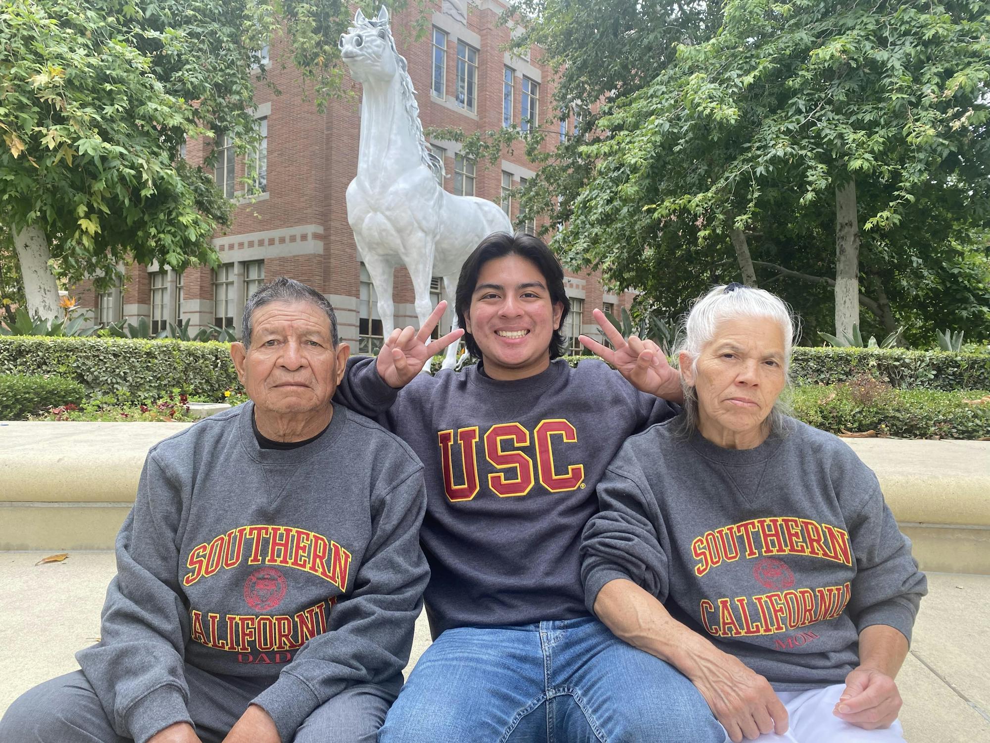A young man sits between his parents, grinning as he holds up two hands in the "Fight on!" handsign. All three wear USC sweatshirts
