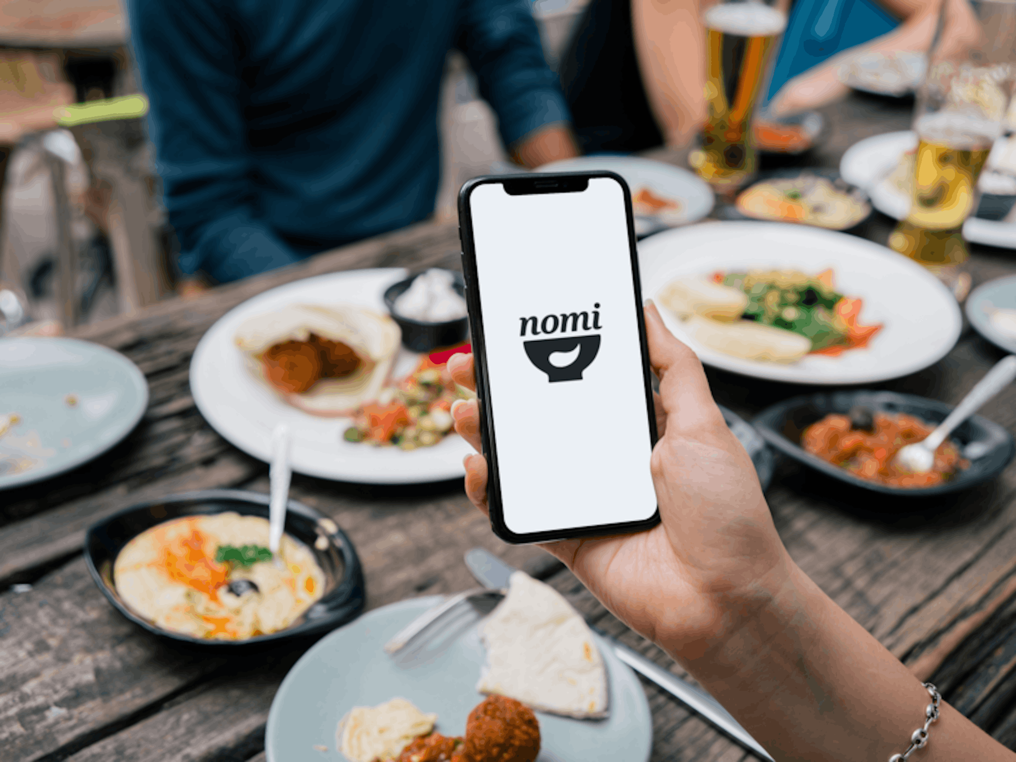 A woman holds her iPhone over a dinner table. The iPhone reads nomi