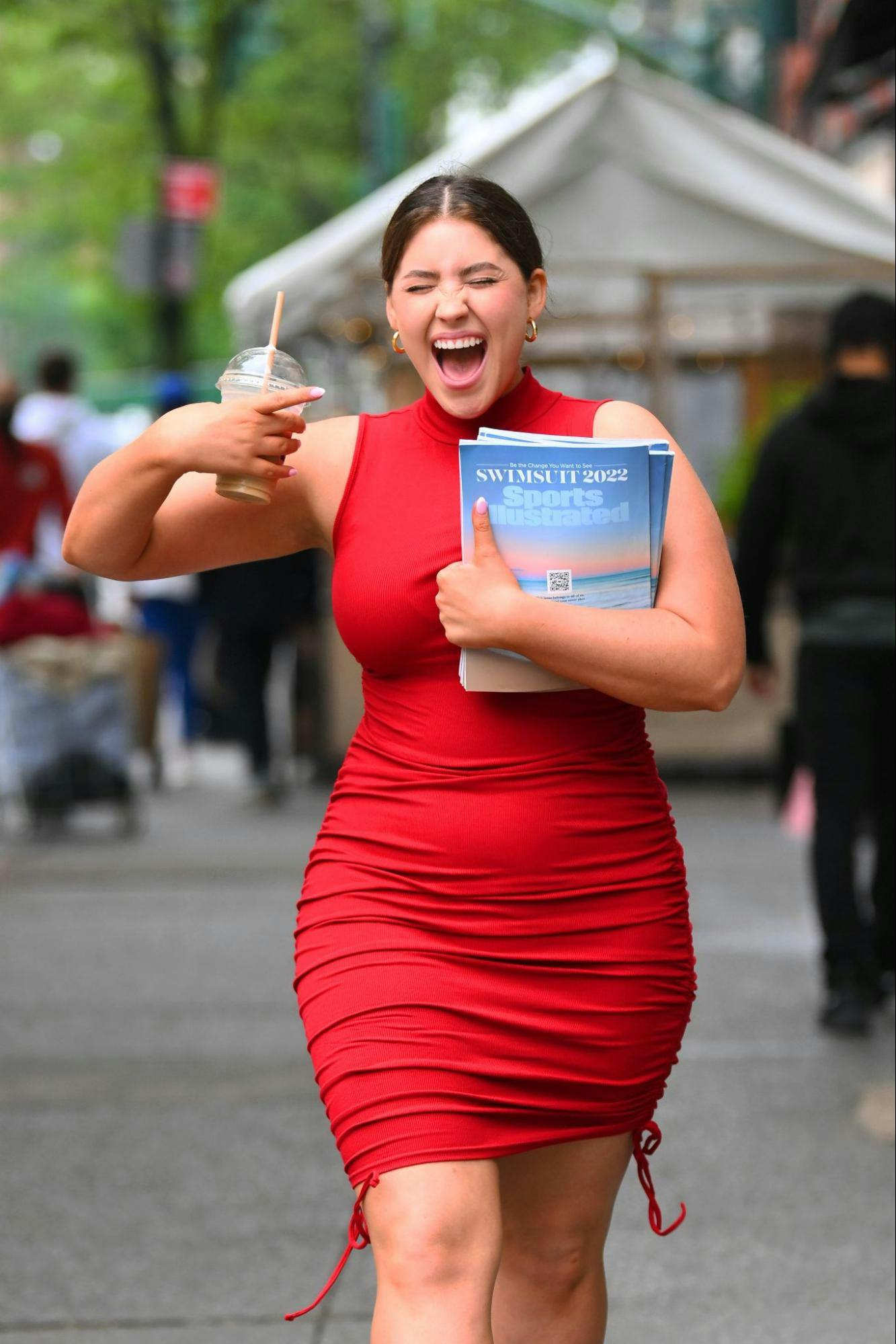 A young woman carrying an iced coffee cries out, grinning, as she holds multiple copies of Sports Illustrated Swimsuit 2022