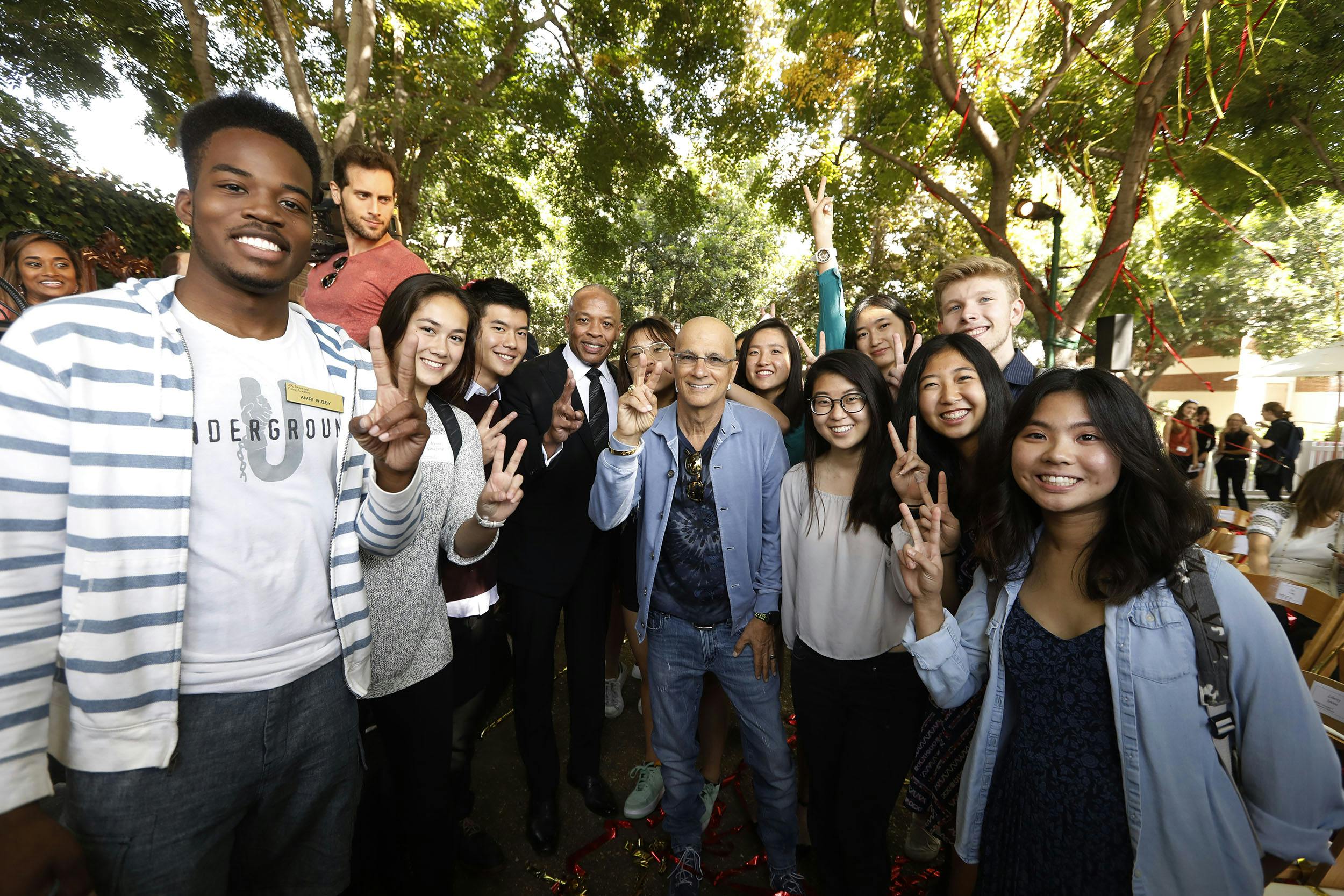 Iovine and Young taking a photo with a group of students. Everyone is holding up a peace sign.