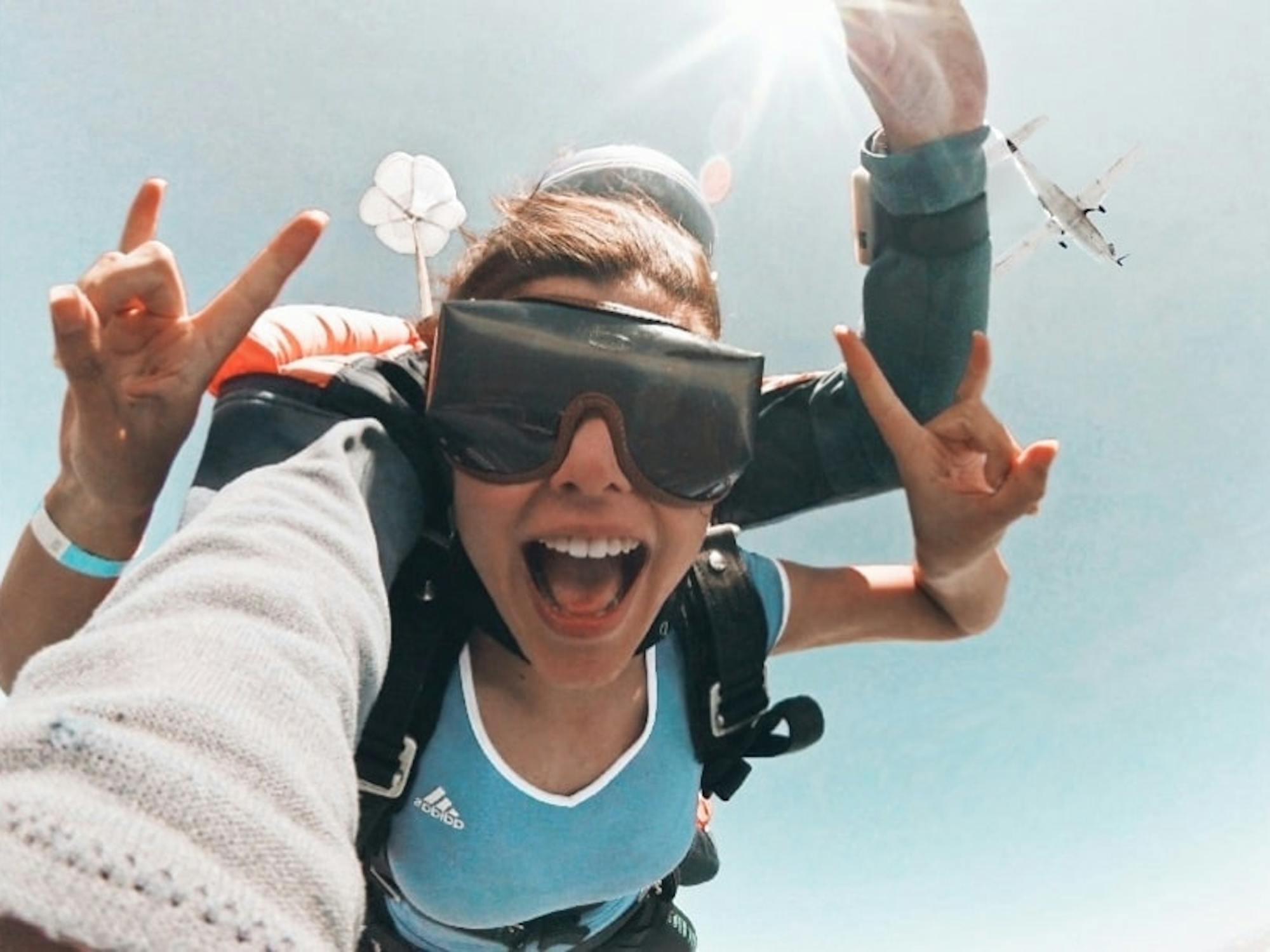 A young woman throws up horns while tandem-skydiving through the air. A plane flies past in the background above