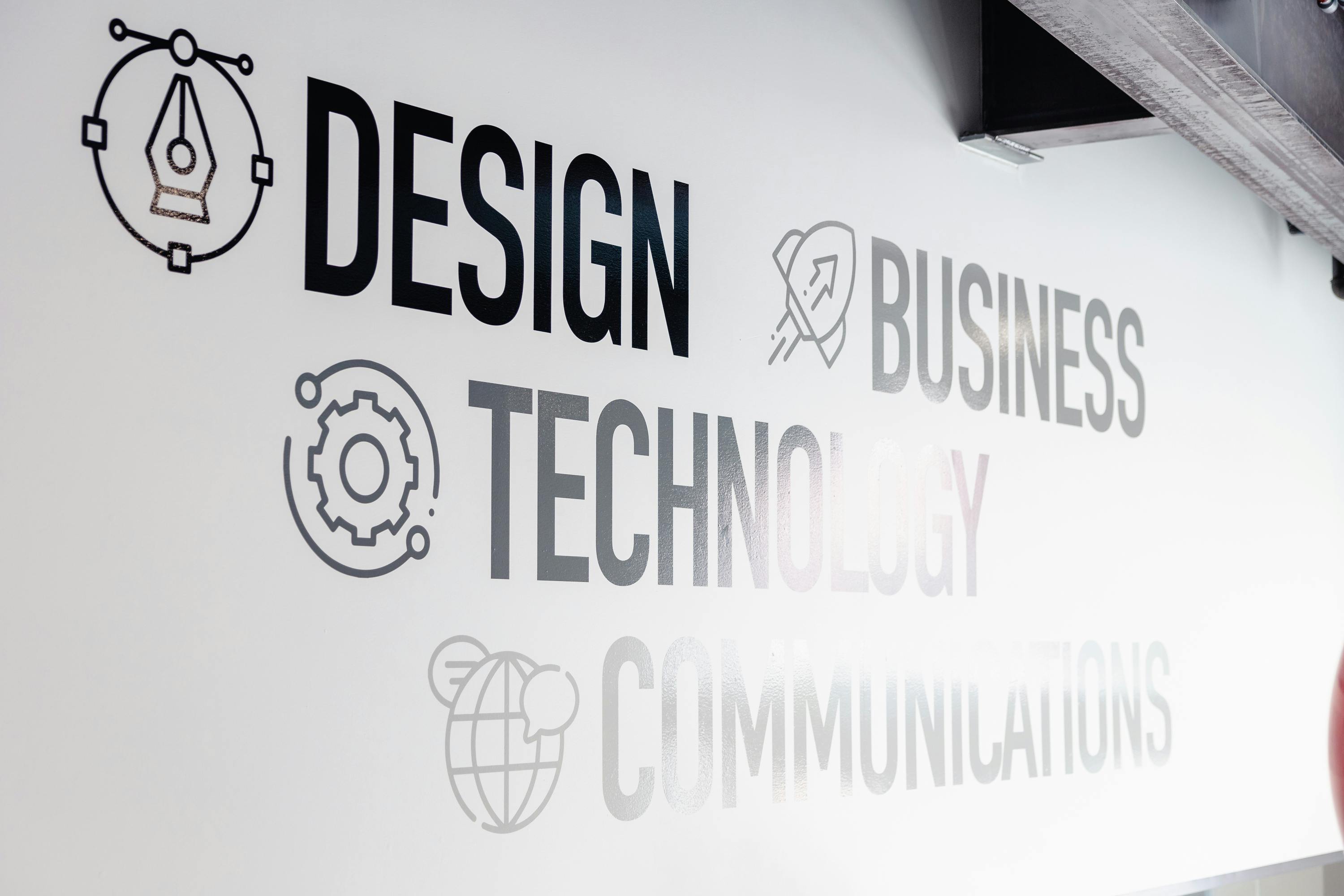 The words "Design, Business, Technology, Communications," each with an icon beside them, are written in bold black font on a white wall