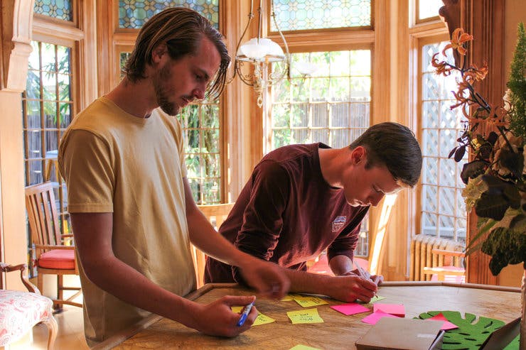 Two young men stand over a table writing on post-it notes