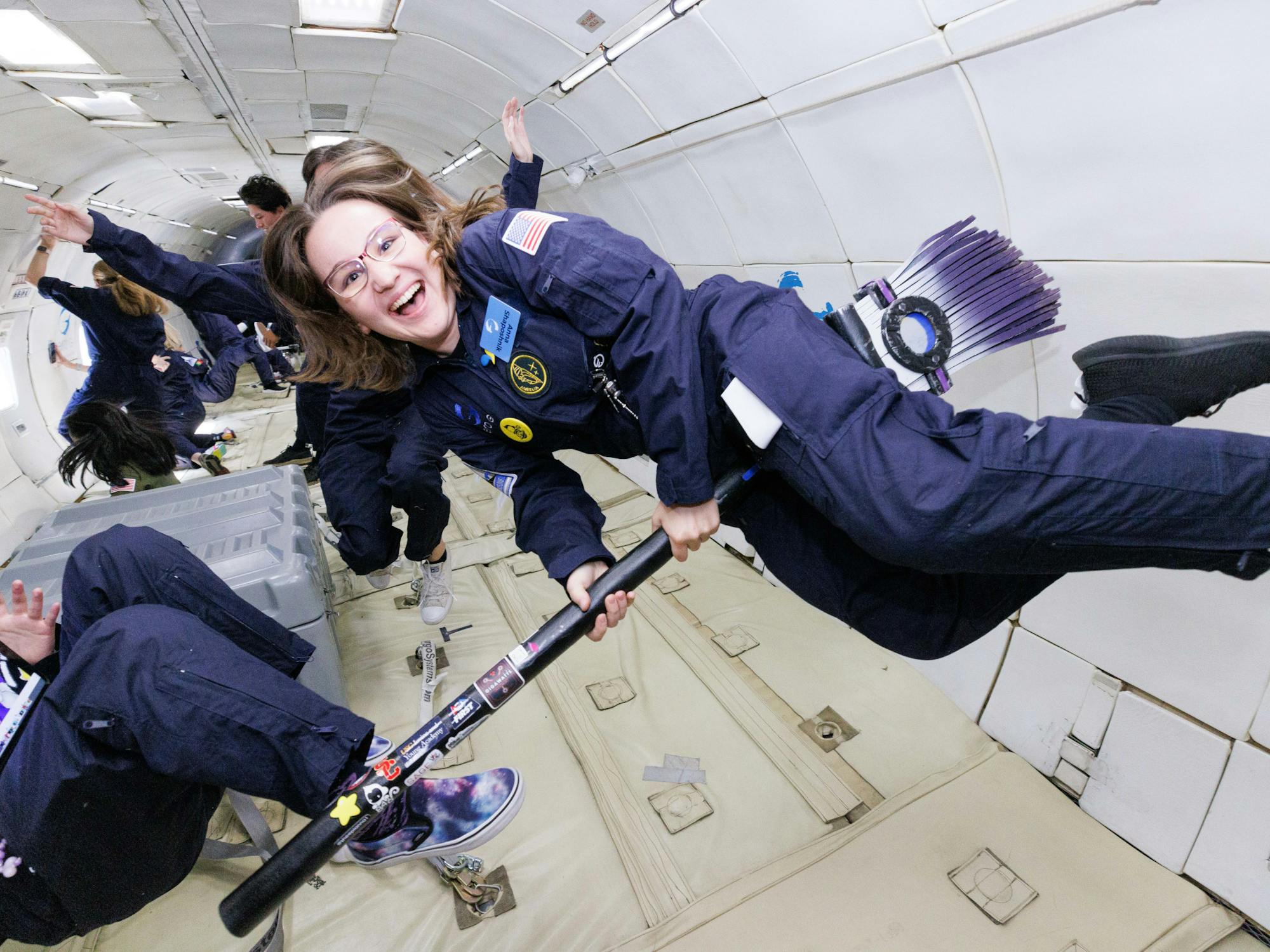 A young woman floats in zero gravity in a flight suit, holding a broomstick