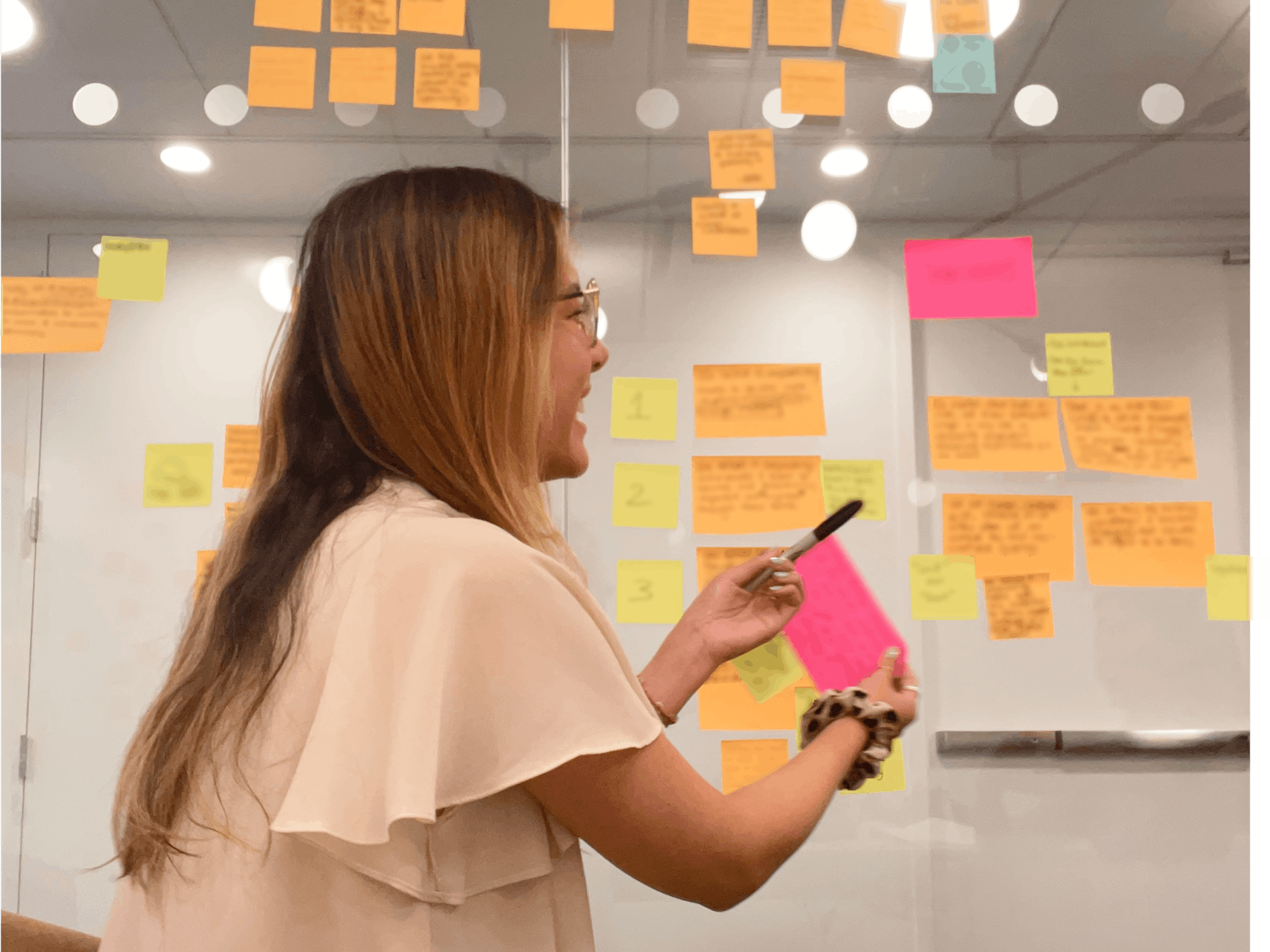 A woman holds sticky notes while standing in front of a board of notes of varying colors 