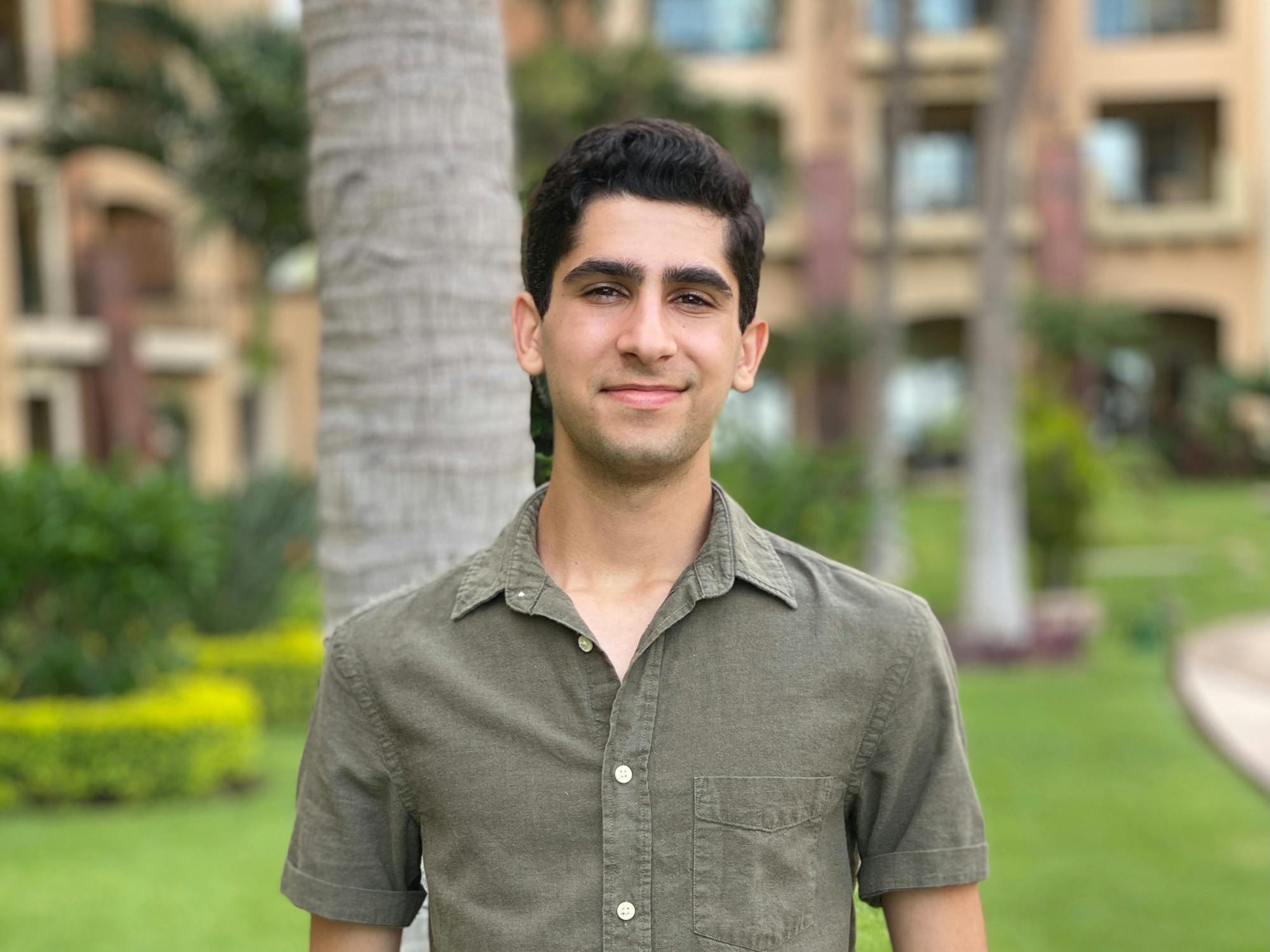 A young man smiles at the camera, posing outdoors in front of a palm tree