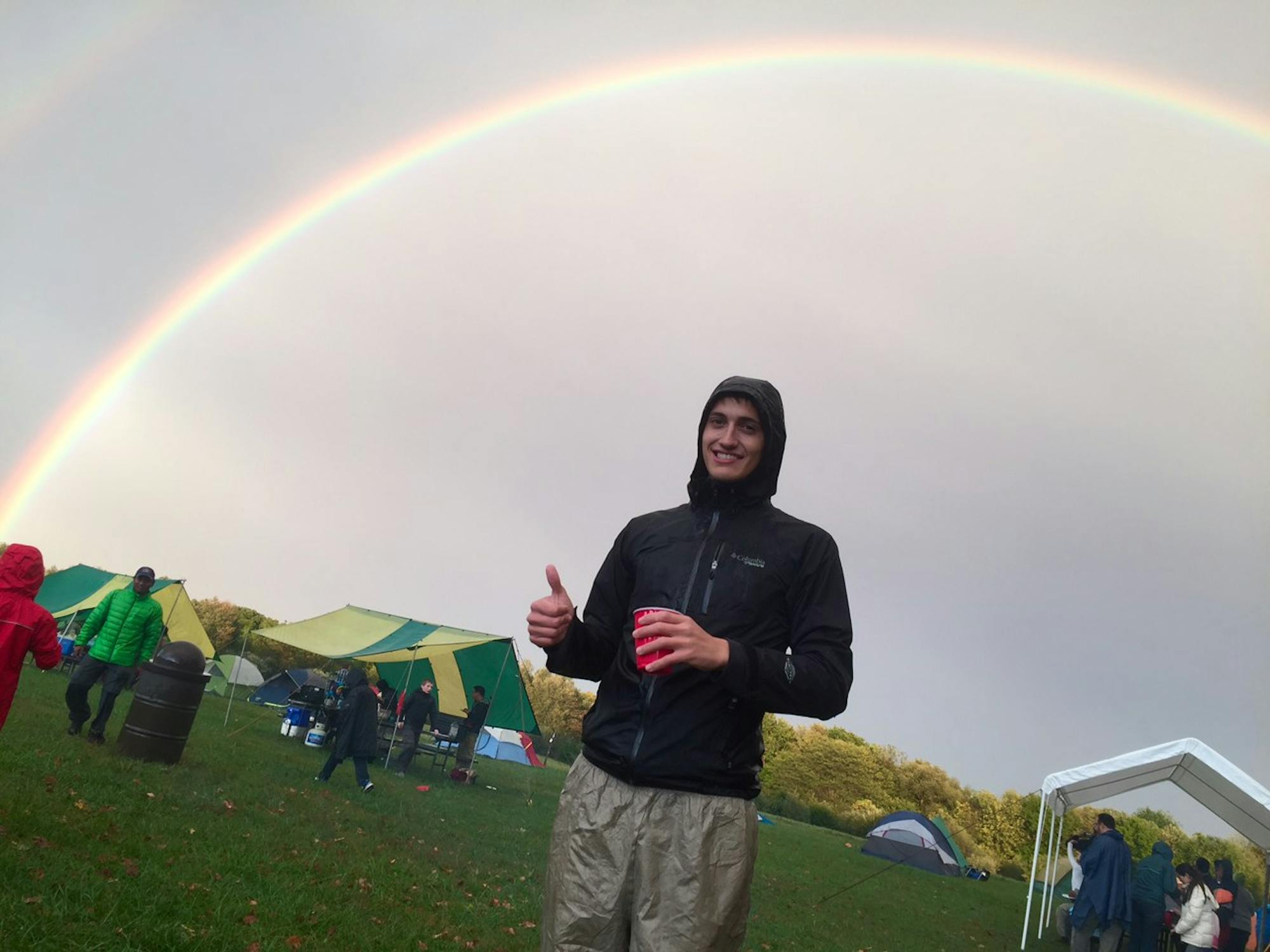 A young man stands outside in a rain jacket, offering a thumbs up. Behind him a full rainbow spans the sky.