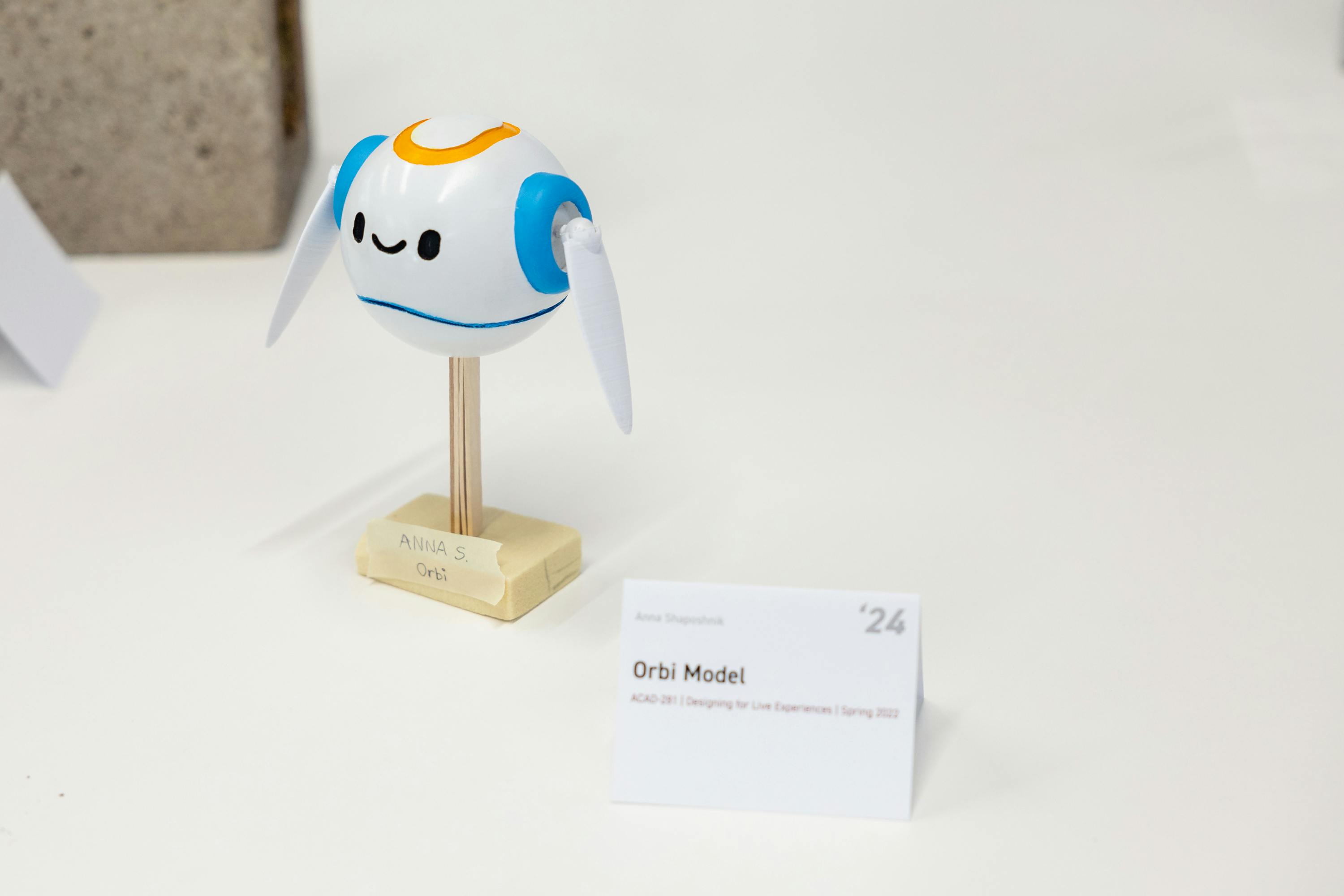 A small toy-like bot labeled "Orbi Model" sits atop a wooden stand