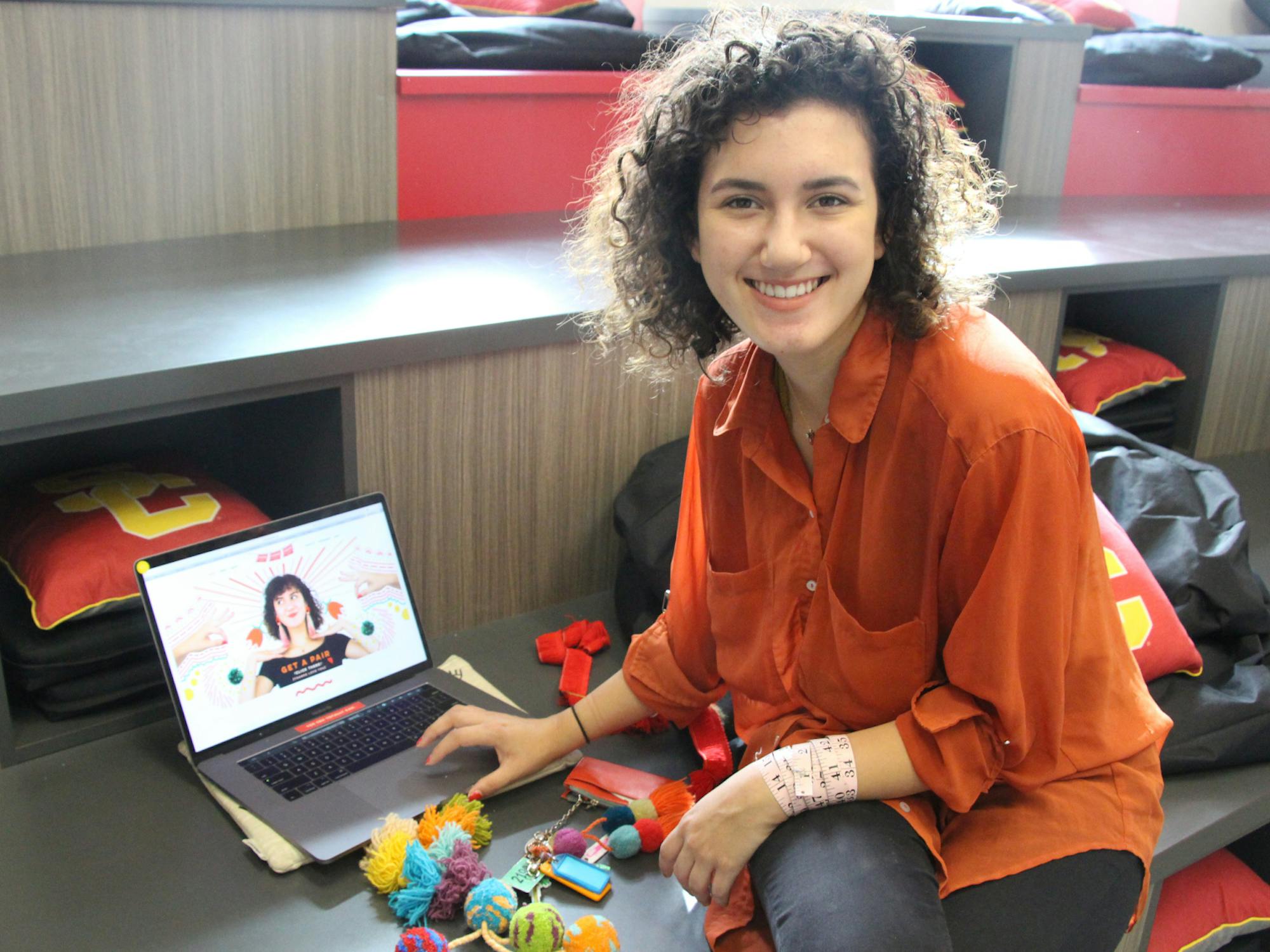 A young woman smiles next to her laptop and a pile of pom poms