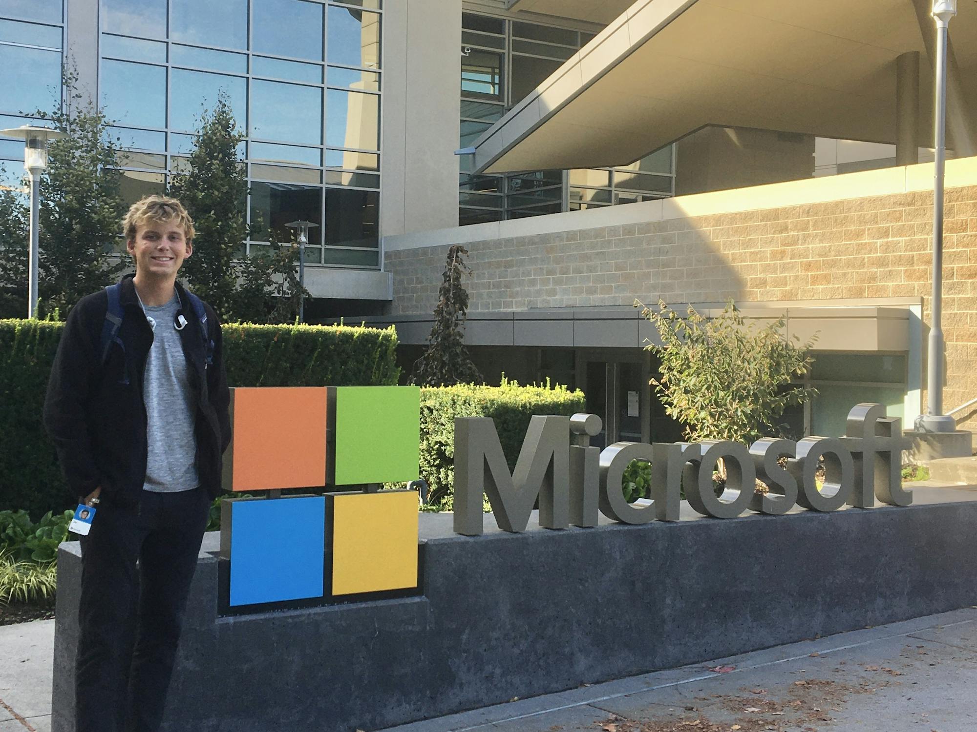 A blonde man in sneakers, a jacket, and a grey t-shirt poses in front of the Microsoft logo