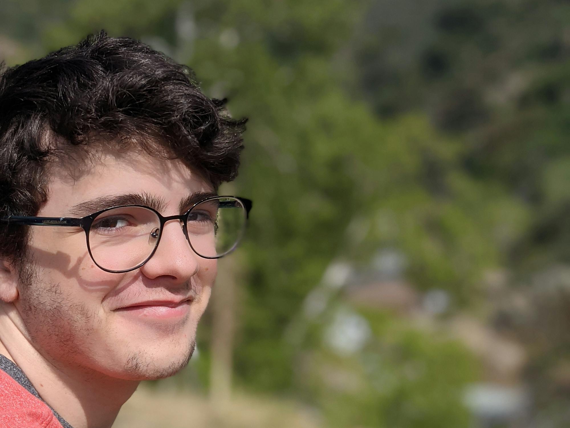 A young man smiles over his shoulder, with trees and mountains in the background