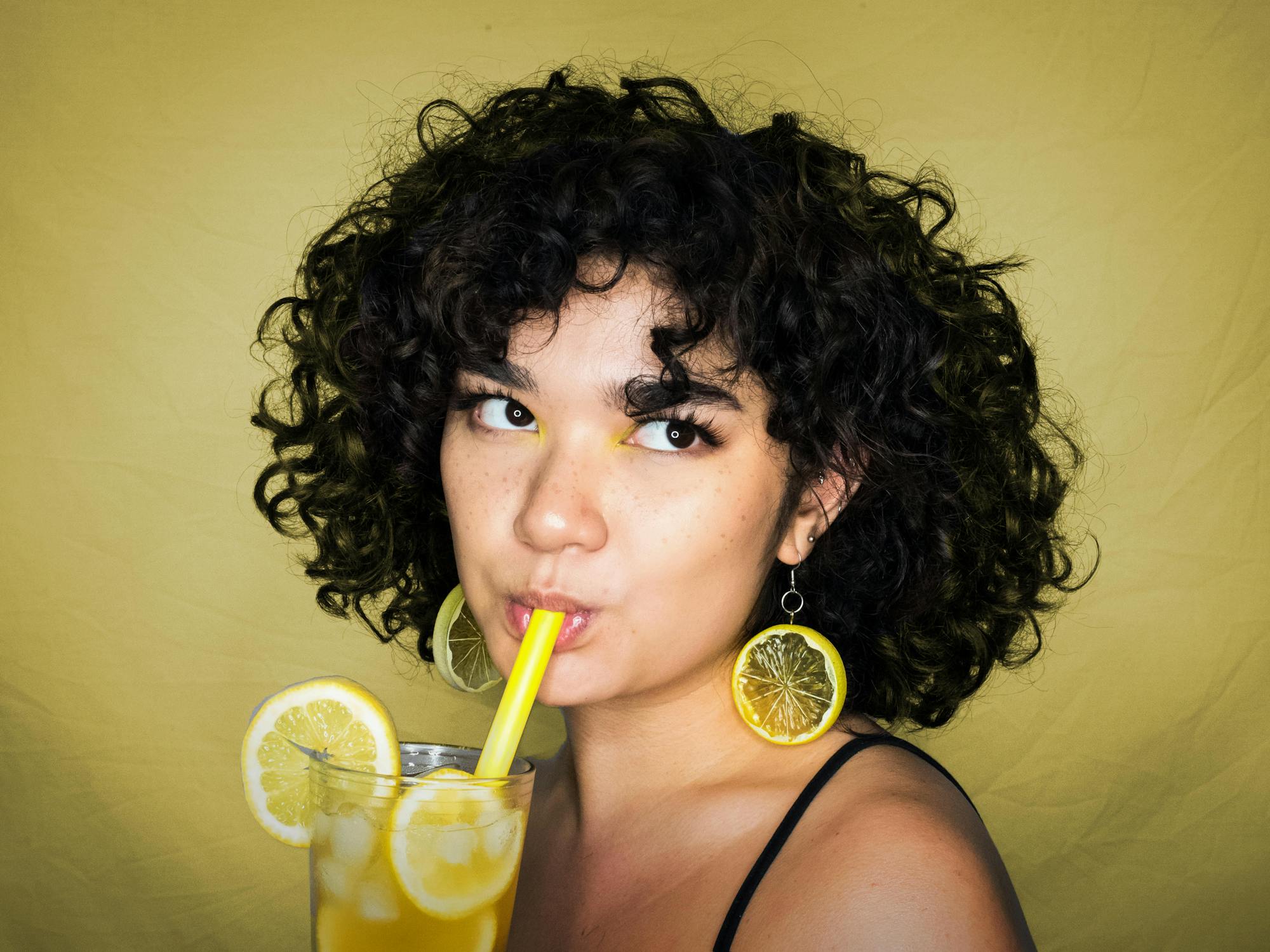 A young woman with curly hair stands in front of a yellow background with lemon-shaped earrings drinking lemonade