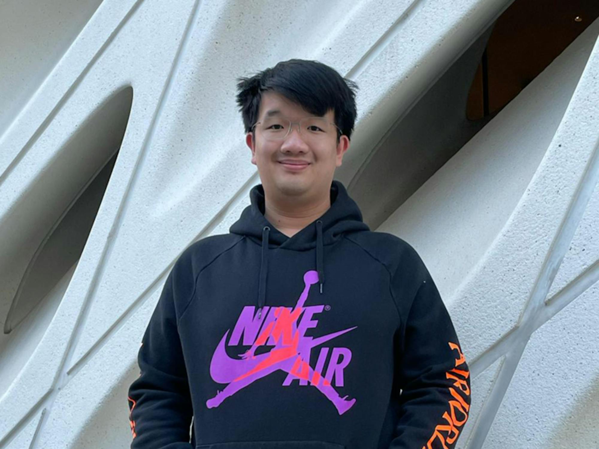 A young man in a hoodie smiles outside an artistically-sculpted building