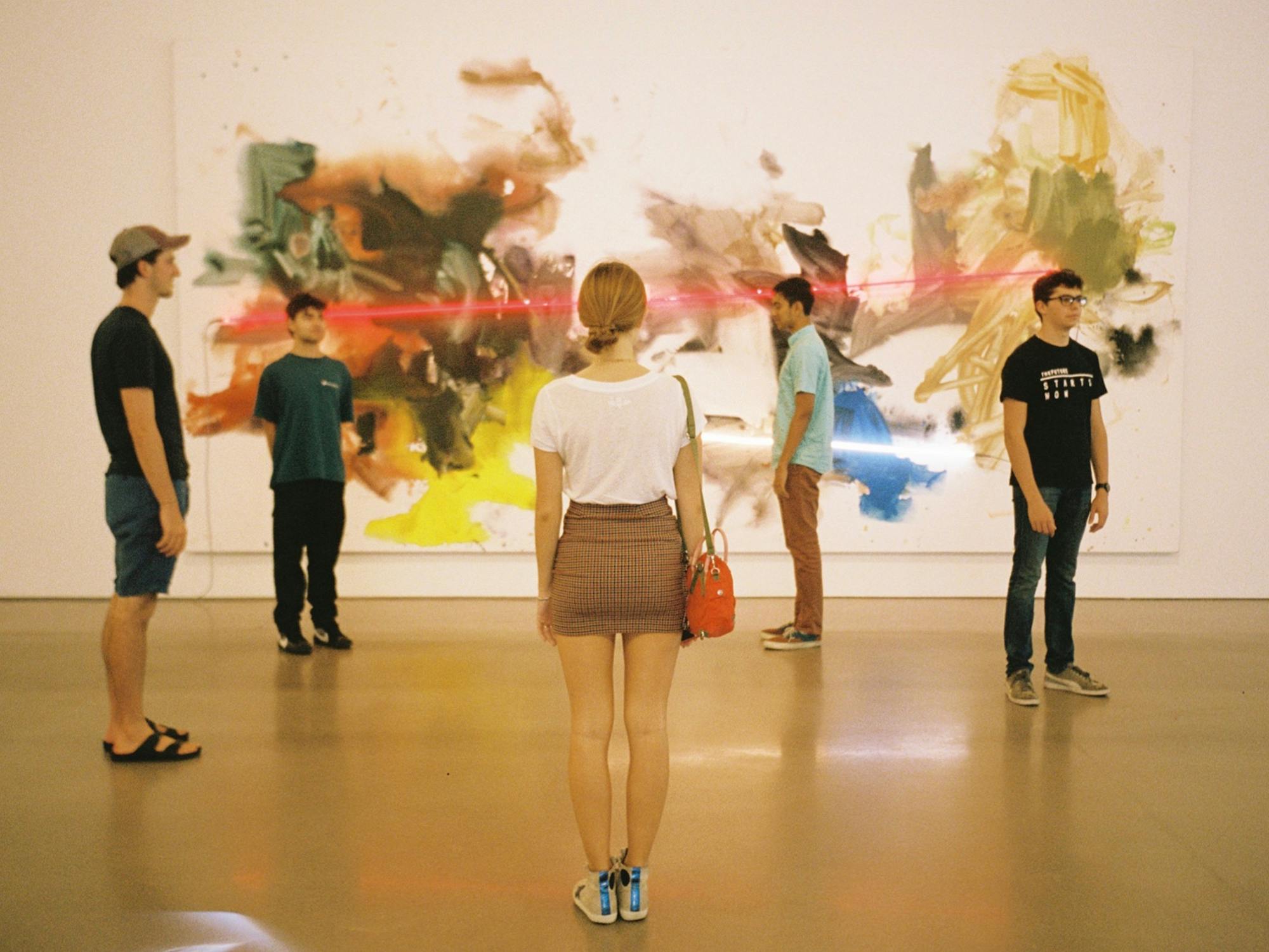 A group of students stand still in a gallery standing stock still. Behind them is an abstract painting