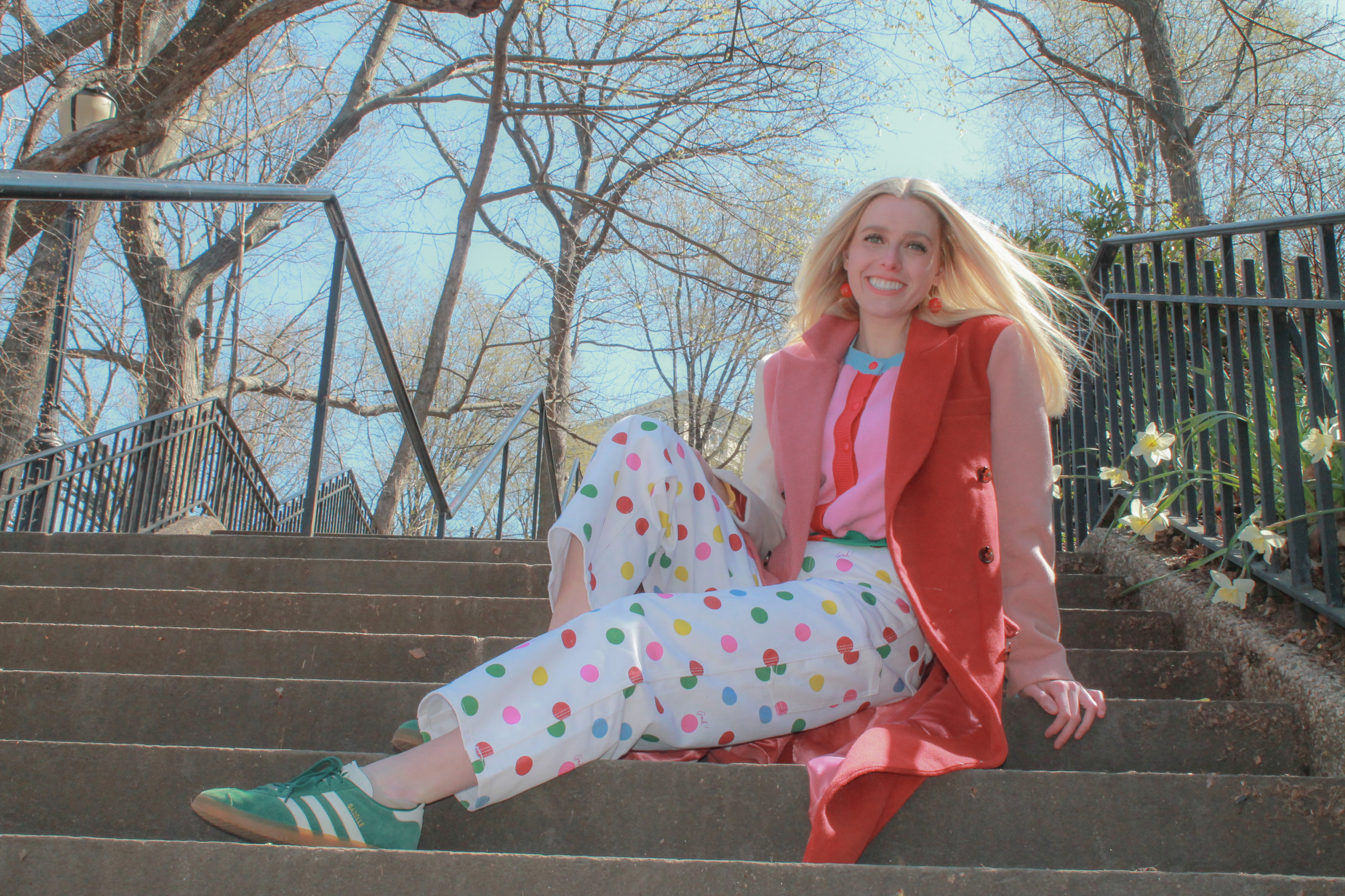 Blonde woman in pink coat and white pants with polkadots sits on steps while smiling. 