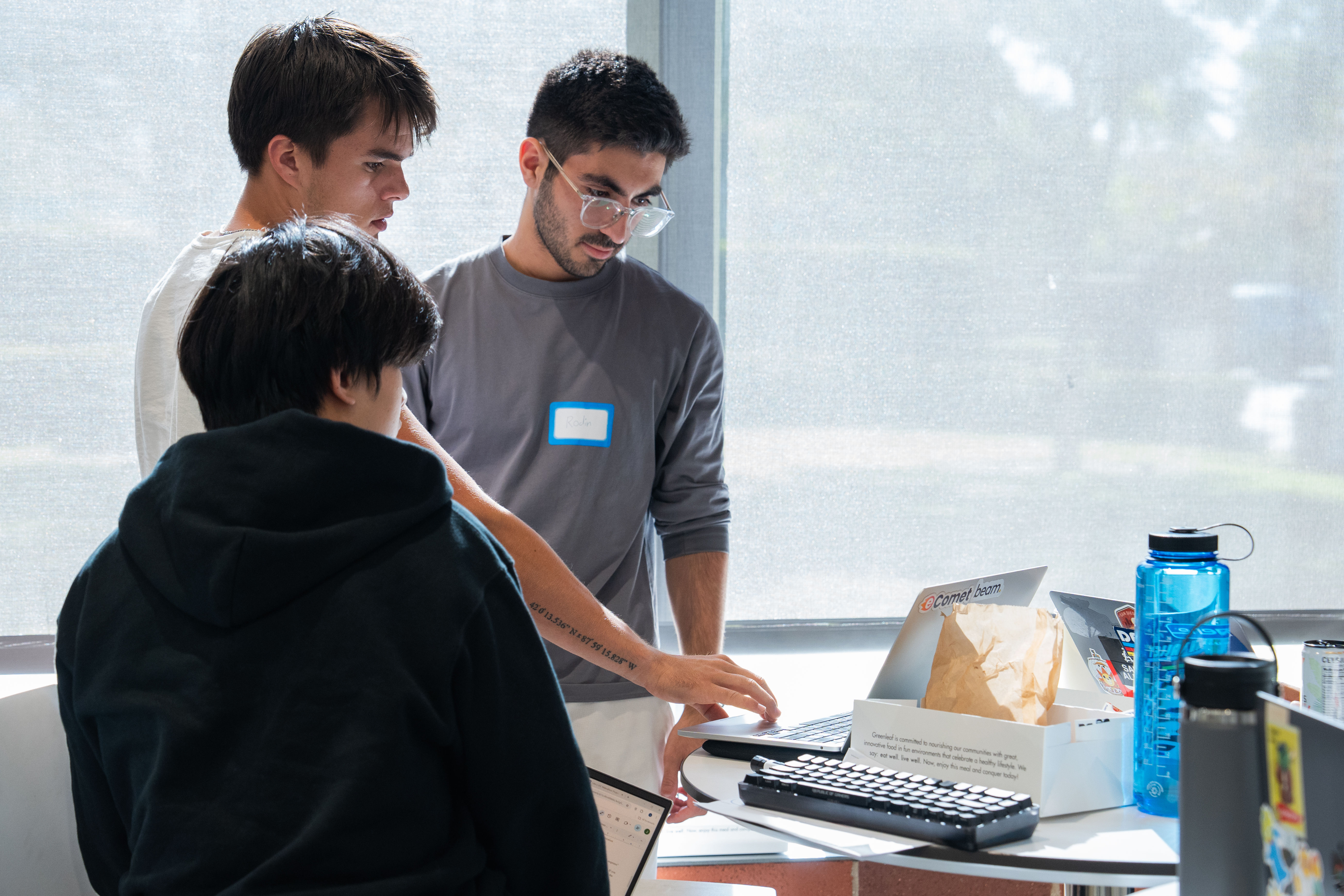 3 male students stand at a high table, all 3 looking at a laptop while one is touching the laptop. 