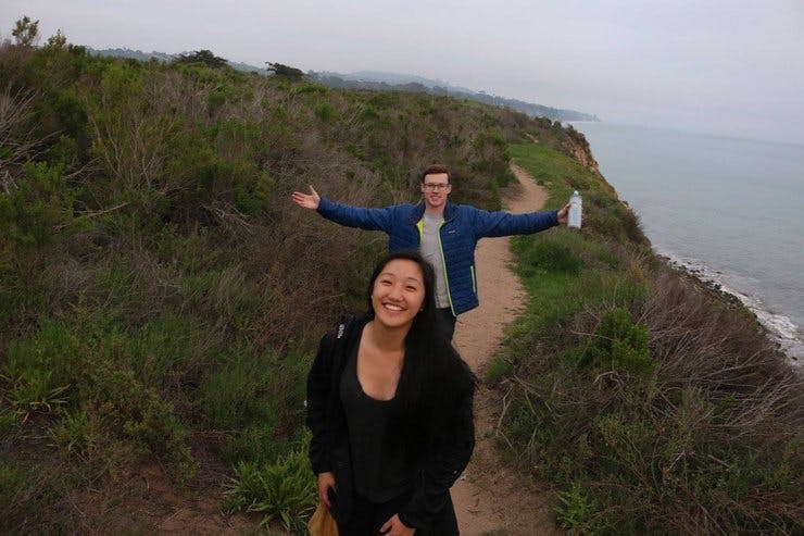 A young woman smiles on an outdoor trail while a young man throws his arms outward behind her