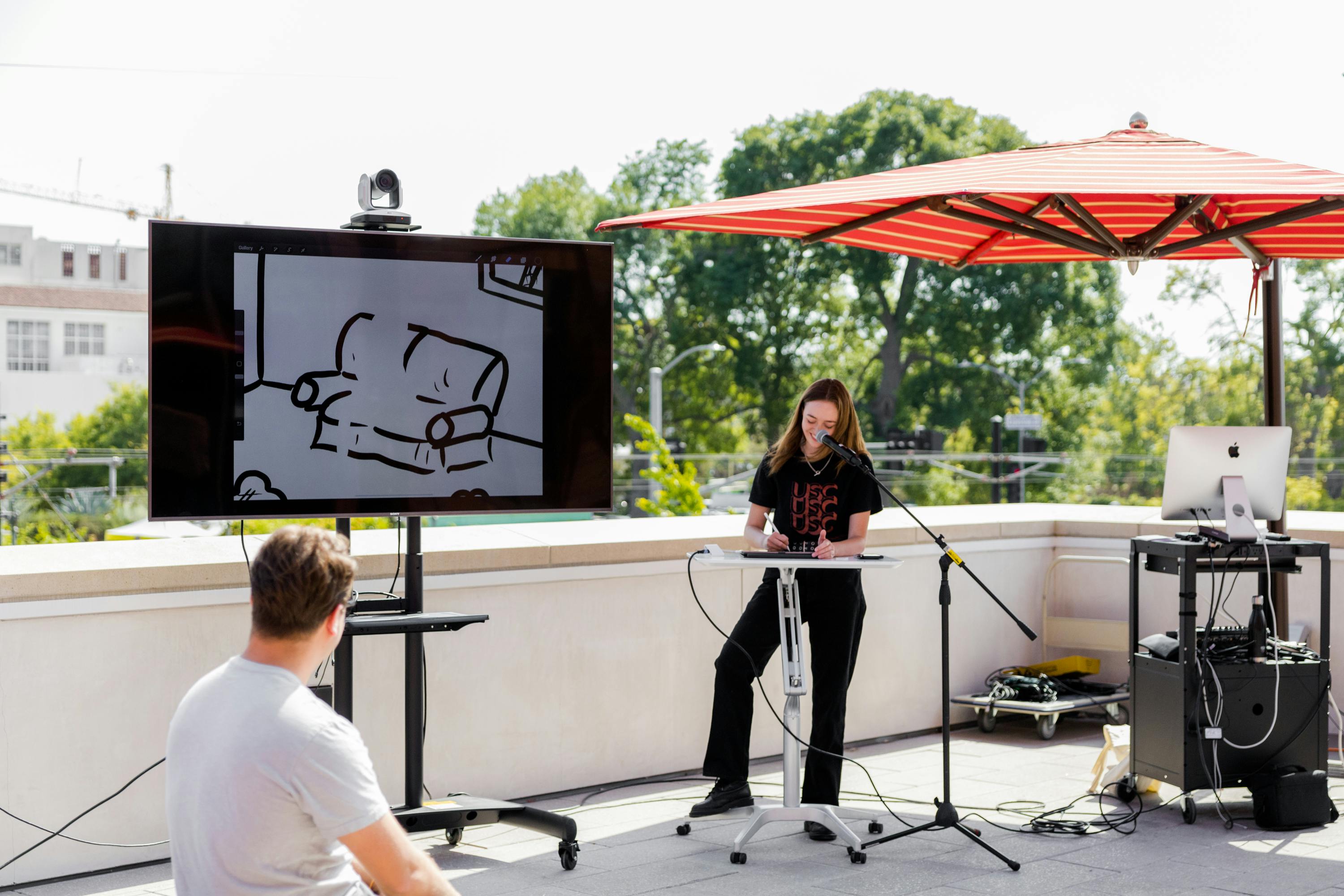 A young woman outdoors at a microphone with a digital tablet projects her results to a TV so an off-camera audience can see it