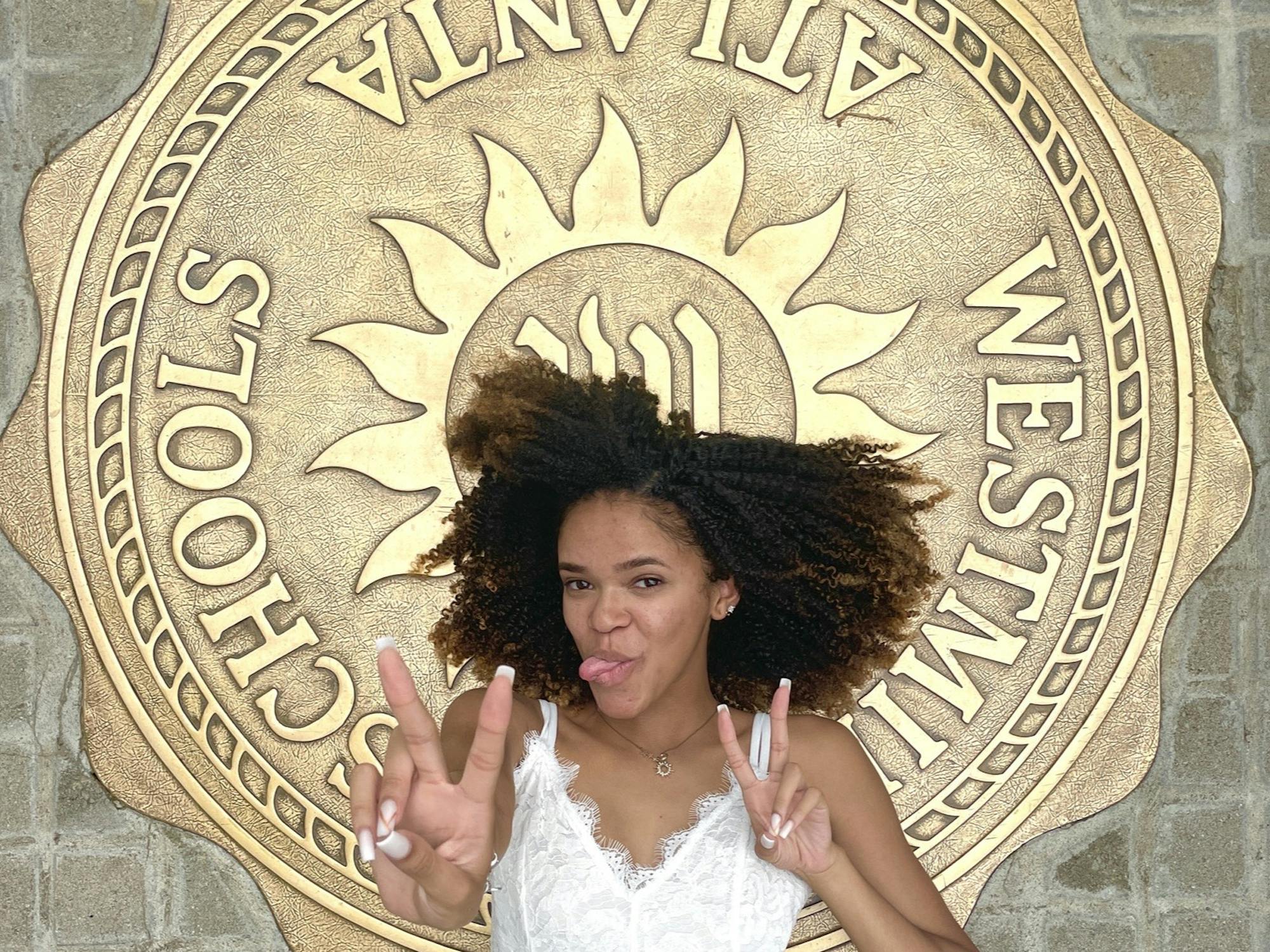 A young woman in a white dress lies on the ground, sticking out her tongue and making a “Fight on!” V with each hand. Over the brick walkway she lies on is a golden metal seal that reads “Westminster Schools Atlanta”
