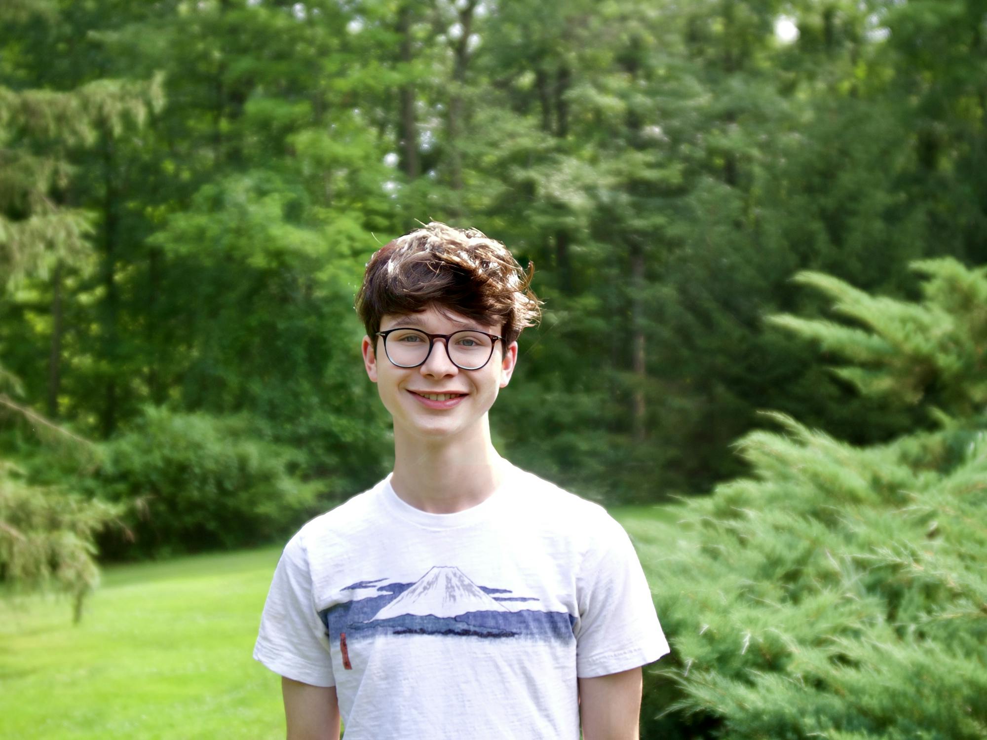 A young man poses outside, smiling before a verdant, forested background.