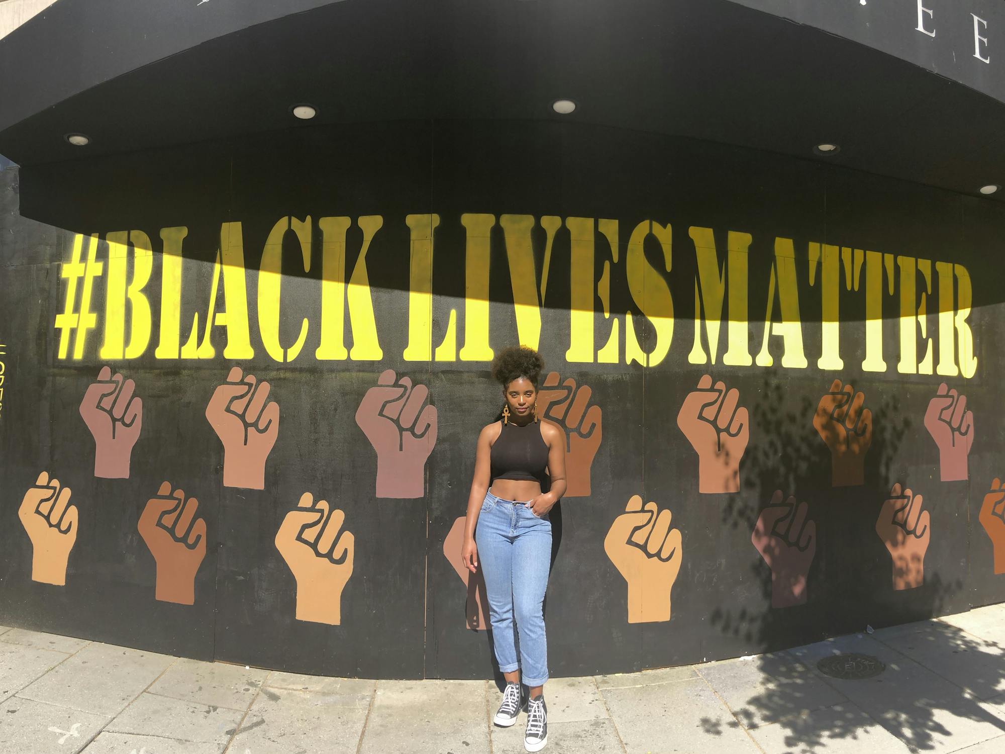 A young woman stands in front of a Black Lives Matter mural with her hand in her jeans pocket and her weight cocked to one hip