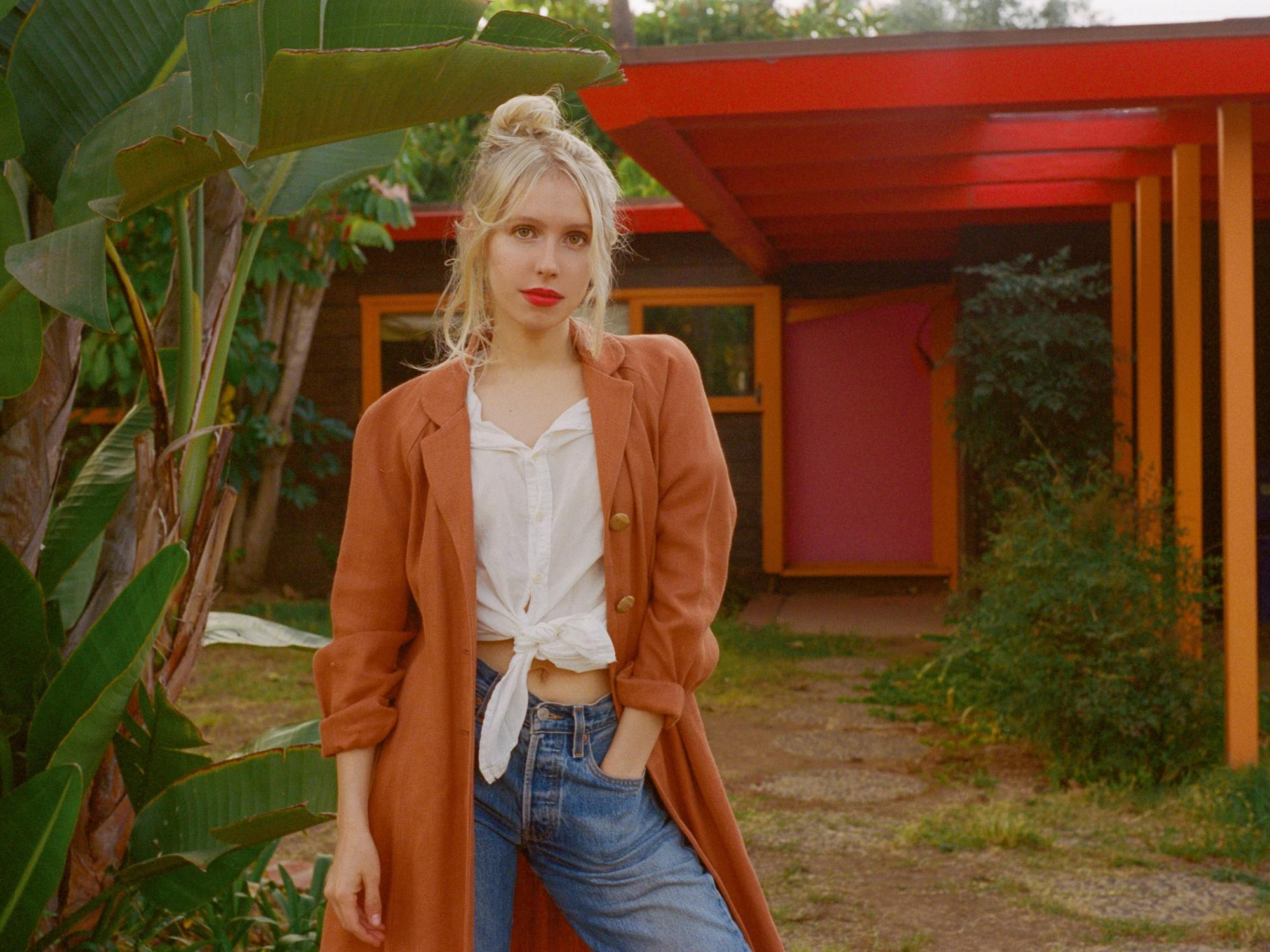 A blonde woman poses with one hand in her pocket. Around her are palms and other greenery, with a single story red building behind her enmeshed in vegetation