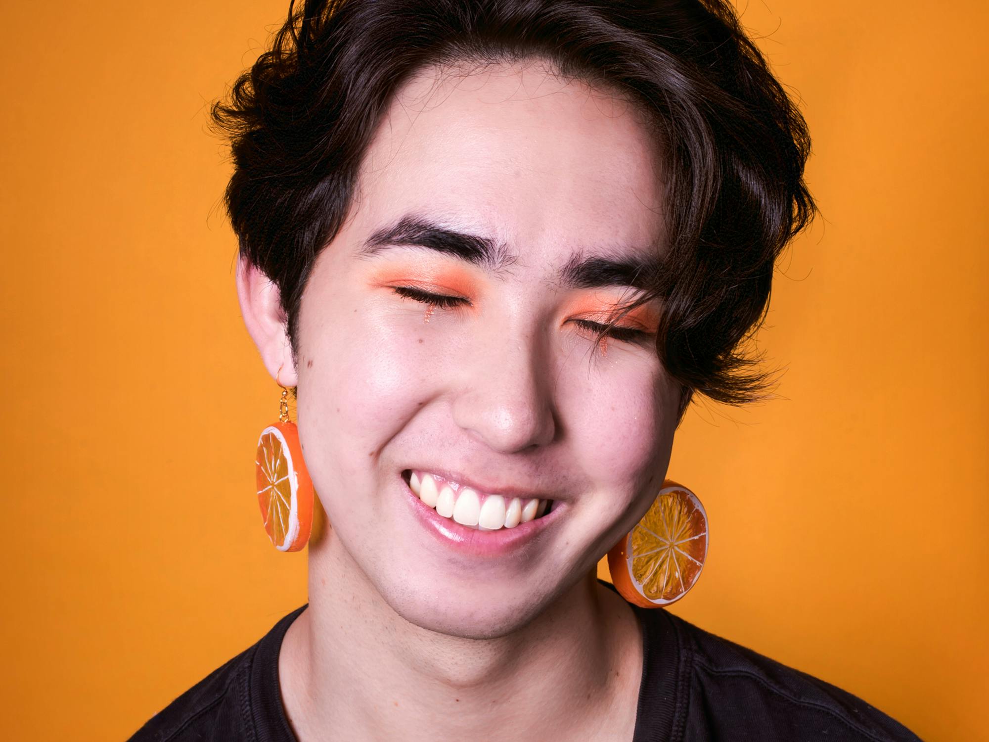 Set against an orange background, a young man with orange earrings and orange eyeshadow smiles with eyes closed