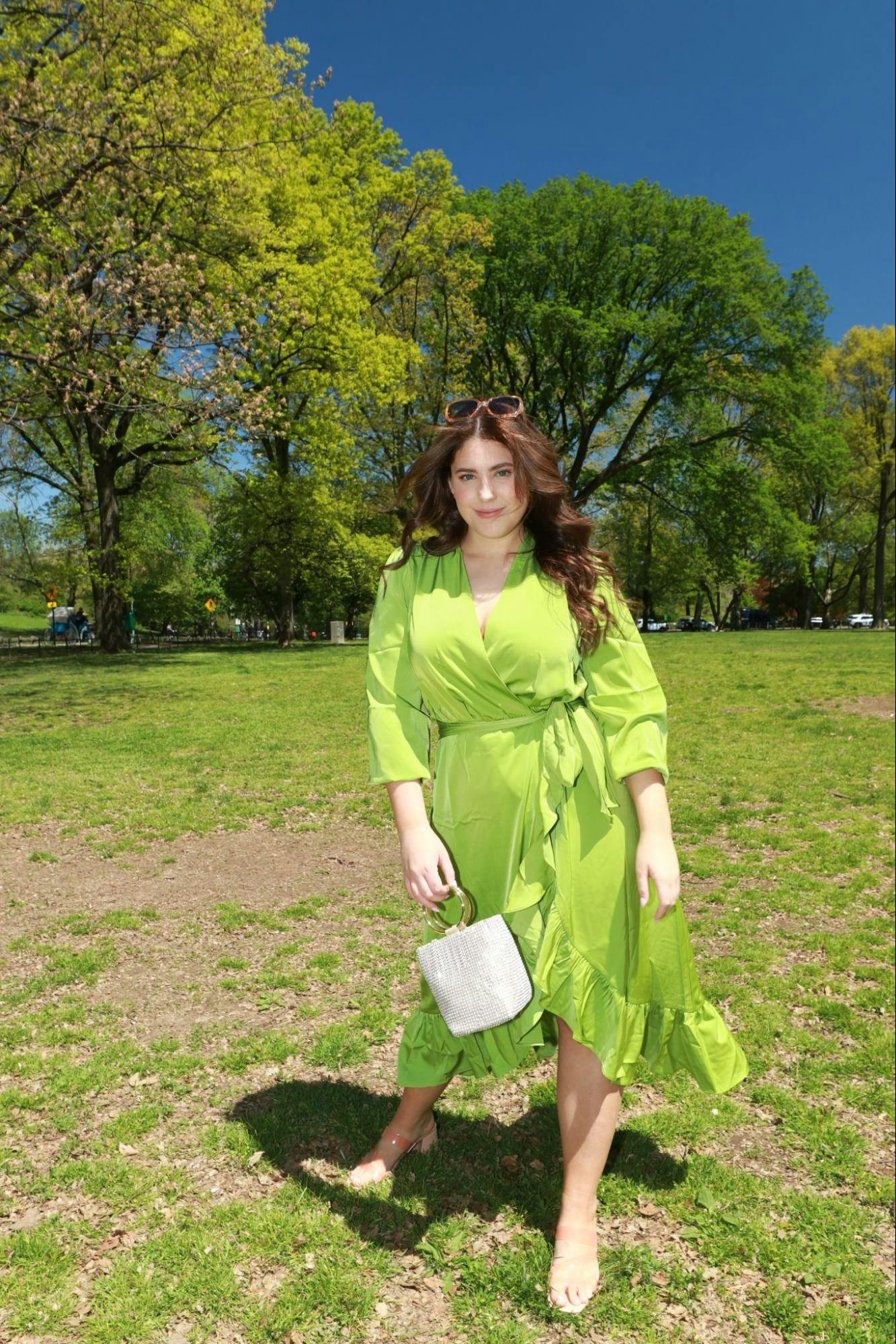 A young woman poses outside on park grass surrounded by green trees. She wears a dress as bright as the plants surrounding her