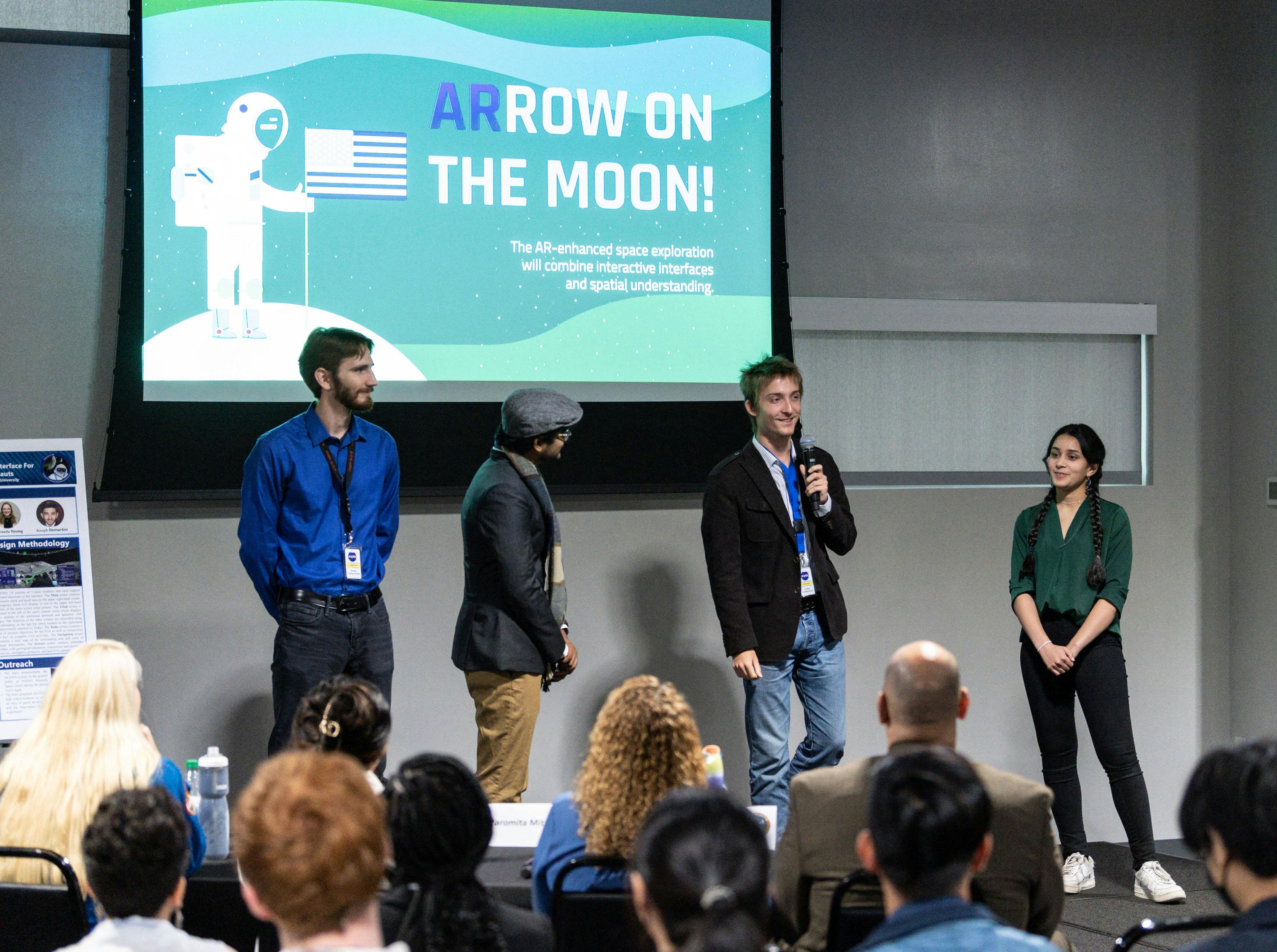 A young man holds a microphone alongside three peers on stage in front of an auditorium audience. Behind them is a slide that reads "ARROW on the Moon: The AR-enhanced space exploration will combine interactive interfaces and spatial understanding"