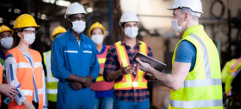 A group of people on a construction site wearing hard hats, masks, and high-visibility vests