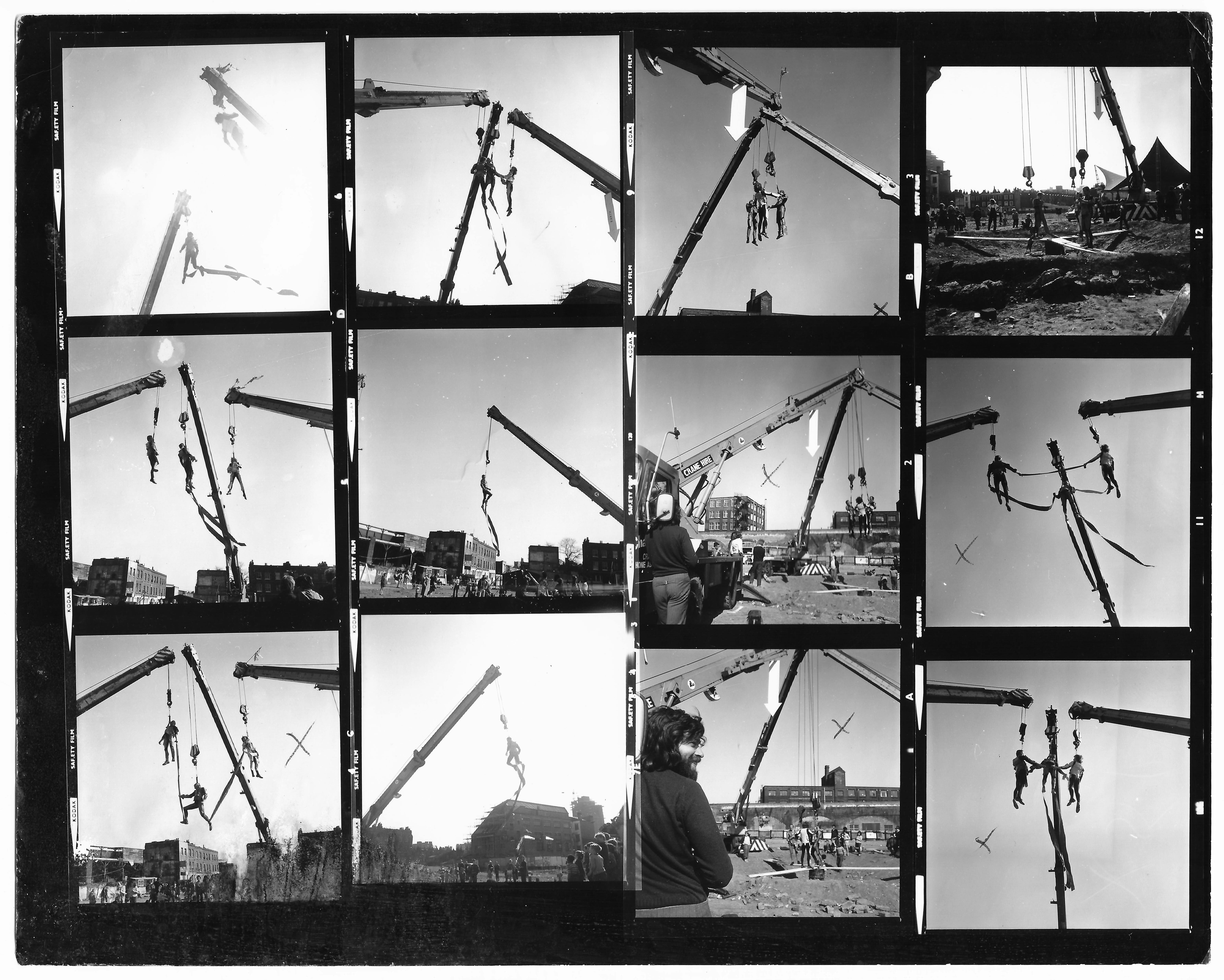 The image is a contact sheet of black-and-white photographs taken during the performance of Leopoldo Maler’s Crane Ballet. The twelve images show three acrobatic performers at various stages of the event. Each performer is harnessed and hoisted from the arm of a crane, occasionally interacting with the others by holding hands or grabbing onto long ribbons hanging from their harnesses. In several images, a large white arrow, pointing down, is seen affixed to the lower portion of one of the crane arms. In a lower image, center-right, the event is seen over the shoulder of Maler, a smiling, bearded man in a sweater.