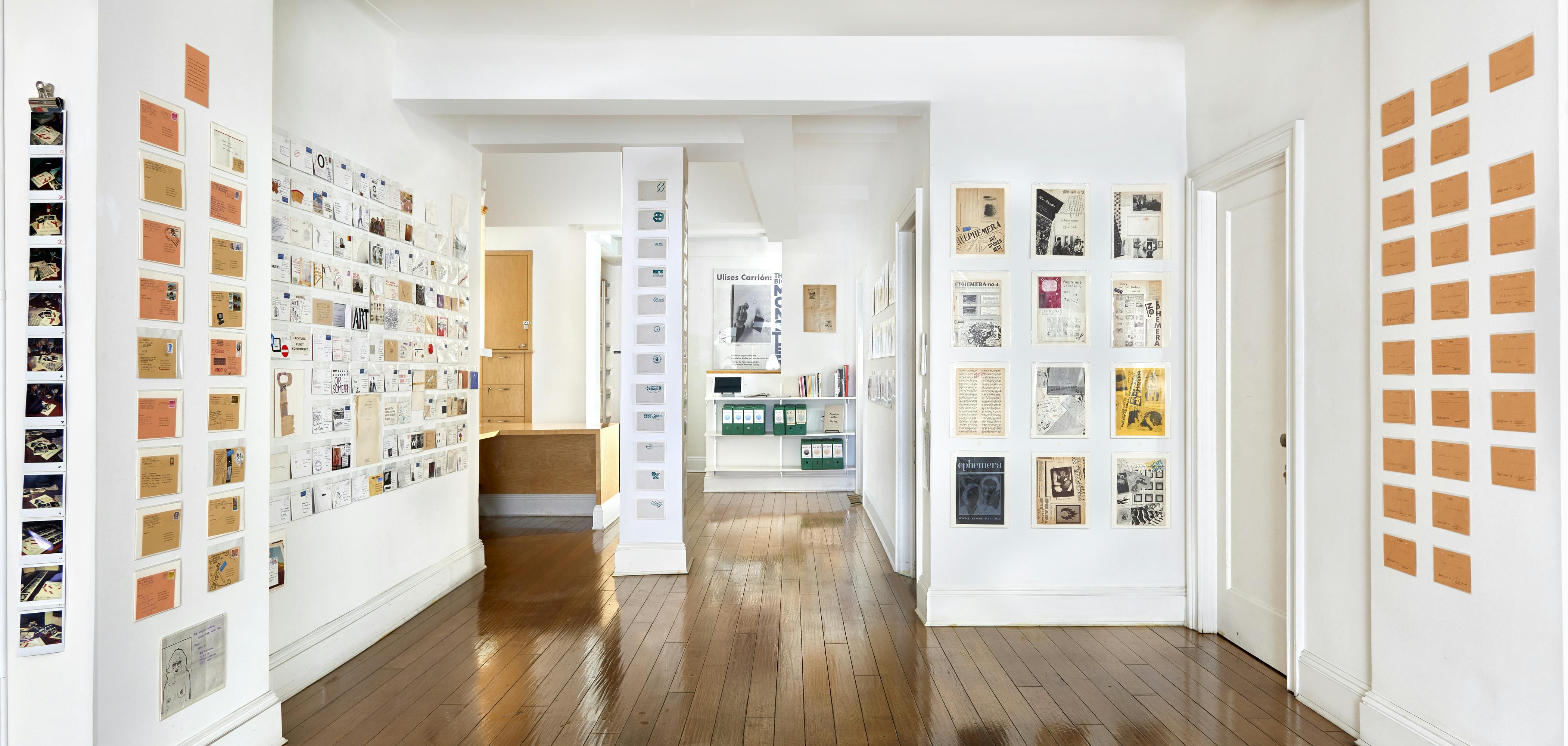 Installation view of Ulises Carrión: The Big Monster exhibition showing envelopes, photographs, and various objects mounted to the gallery walls.