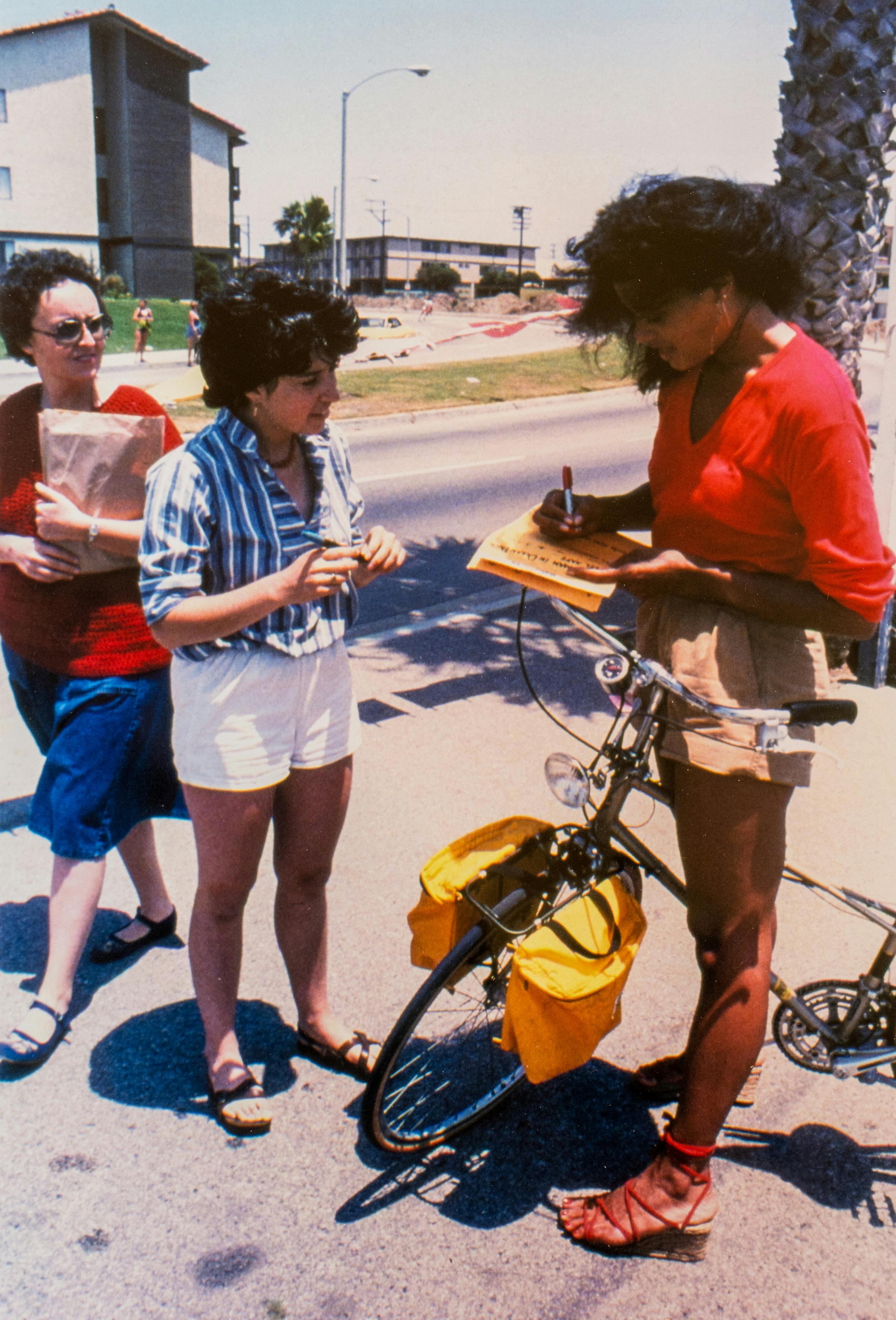 A color photograph of three woman standing on a sidewalk. One woman stands with her bicycle and writes on a piece of paper that she has balanced on her handlebar. Another woman, holding a pen, watches her and seems to be waiting for her to finish writing. A third woman, holding a brown paper bag, walks past, seeming to glance hurriedly at the woman filling out the form.