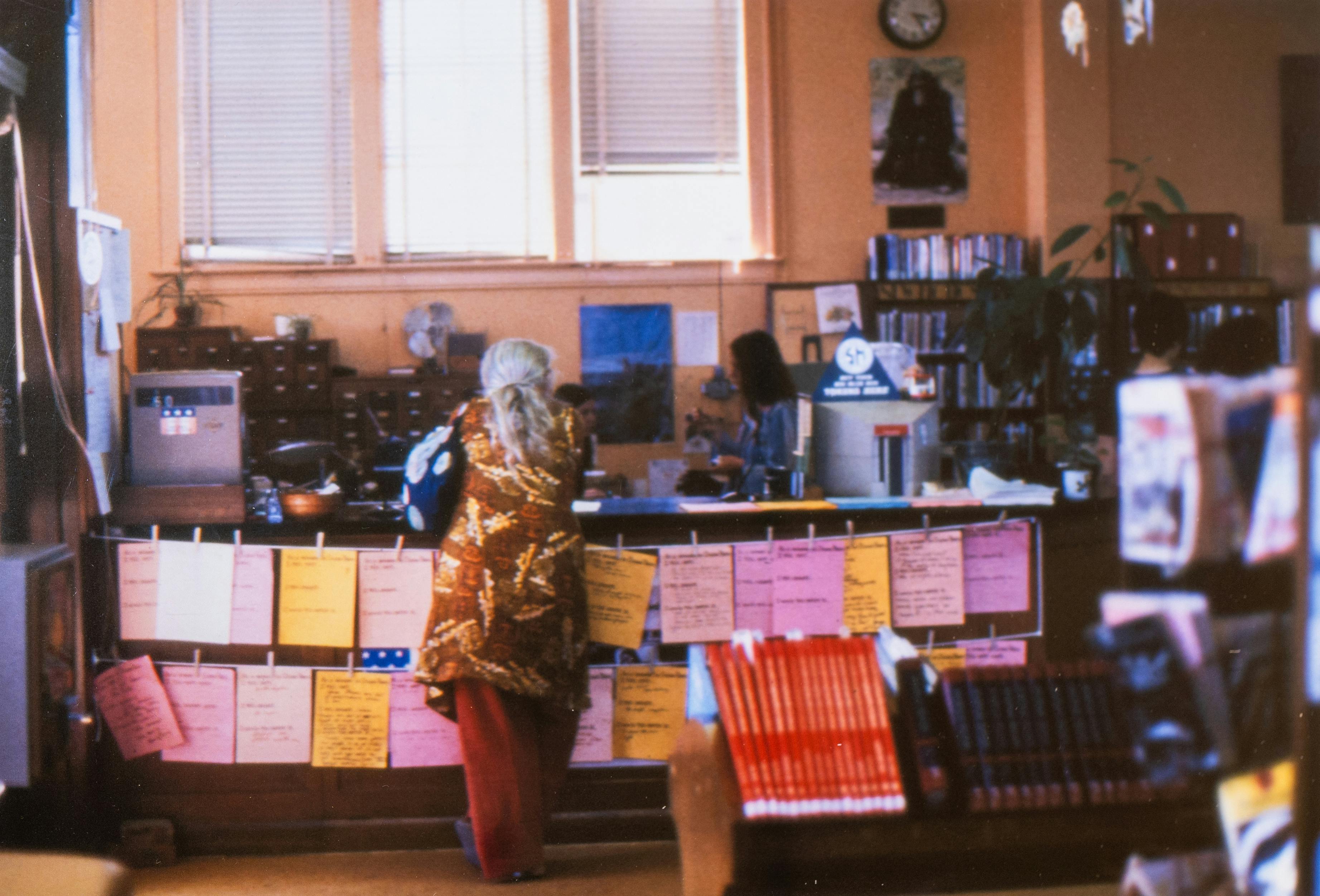 A color photograph of the interior of a bookstore, in which we can see a busy countertop with register, catalog card cases, and inventory. Suspended in two rows on the front face of the checkout counter are sheets of pink and orange paper with handwritten text hung from two clotheslines. A woman leans against the countertop, speaking to an attendant behind the counter. Her body partially obscures the sheets of paper.