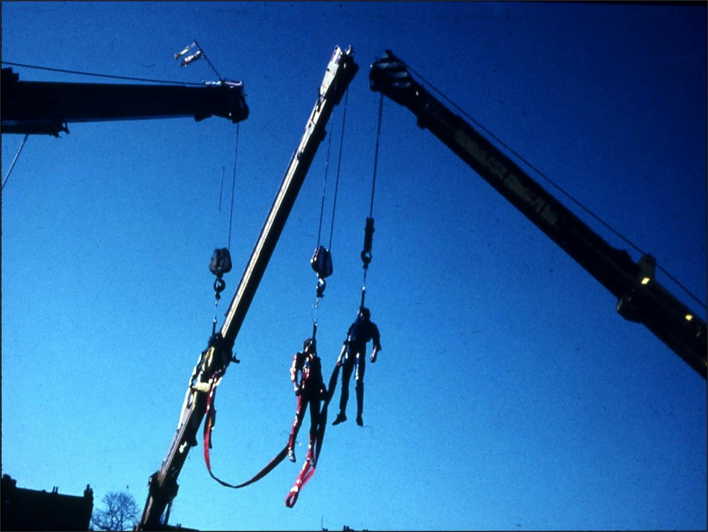 A blue-tinted photograph in landscape orientation. Taken from the ground, the photo shows three performers suspended in the air from harnesses attached to the arms of three mobile cranes. The performers hang loosely, connected to one another by thick bands of red ribbon trailing down from their harnesses.