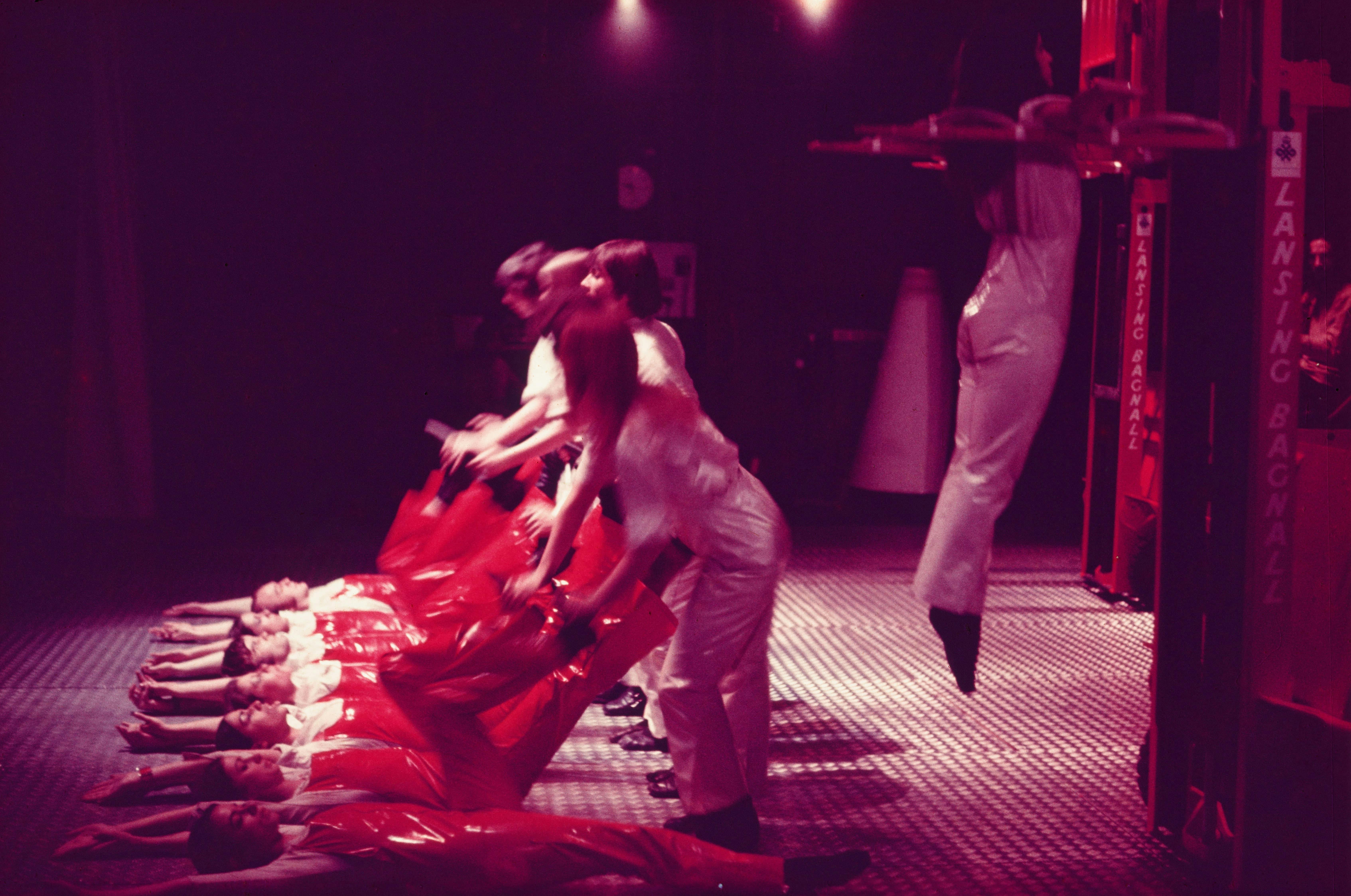 The image shows a color photograph taken during the 1970 performance of Leopoldo Maler’s X-IT2 at The Place in London. Performers wearing red vinyl costumes lie in a line on an industrial floor, their arms above their heads and their legs kicked up against a corresponding line of standing performers in white, who are holding their feet. Behind this group, another performer in white is suspended a few feet off the ground by the arms of a forklift. The performer has his arms outstretched and his legs held together in a pose evocative of crucifixion.