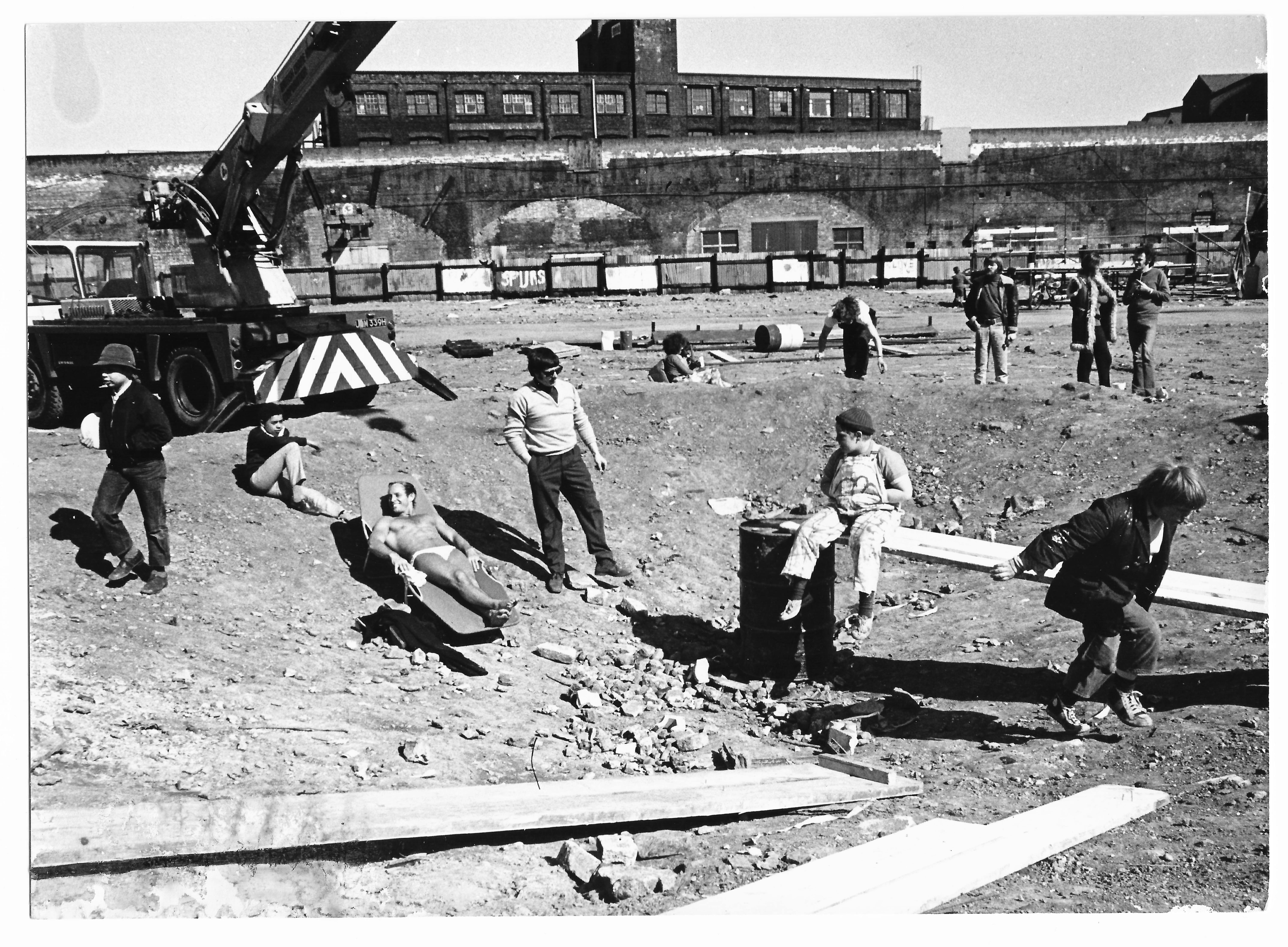 The image shows a black-and-white photograph taken during the performance or rehearsal of Leopoldo Maler’s Crane Ballet. On an unpaved construction site where a circular pit has been dug, several boys run around or sit on a plank of wood, which is propped up against an oil drum near the center of the image. To the left, a smiling man in a white speedo sunbathes on a recliner. Behind him, the front portion of a mobile crane is visible in the background, its arm extended upward out of the image’s frame.