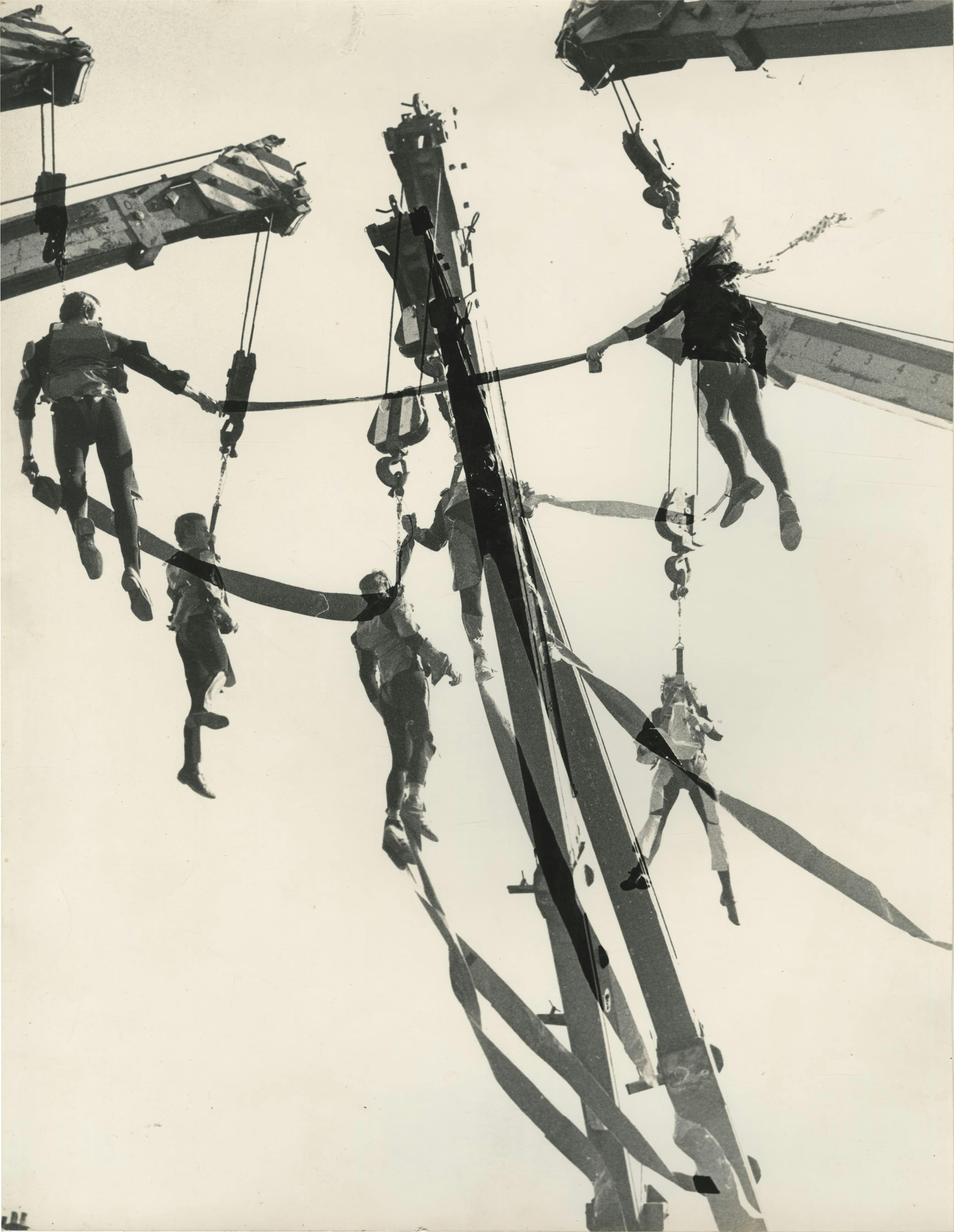 The image shows a black-and-white photomontage taken during the performance or rehearsal of Leopoldo Maler’s Crane Ballet. In the composite image, the arms of six cranes enter the white space of the sky from various angles—two from the upper left, two from the upper right, and two from directly below—and overlap in the center of the image. From each of these crane arms, a harnessed acrobatic performer hangs, many of them trailing or holding onto thick ribbons that hang from their costumes and connect them to one another.