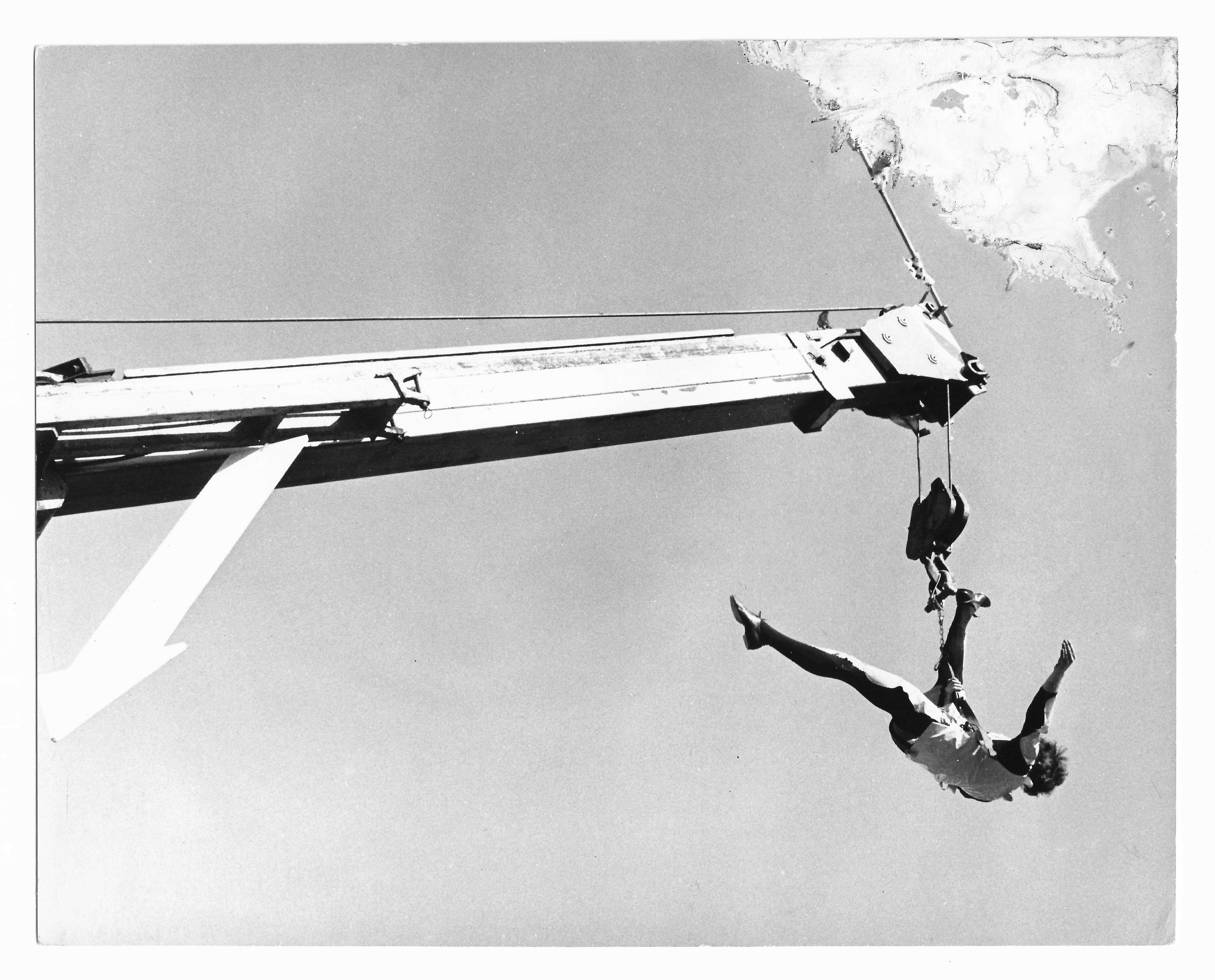 The image shows a black-and-white photograph taken during the performance or rehearsal of Leopoldo Maler’s Crane Ballet. The image, shot from below, shows a mechanical crane arm extending vertically from the bottom of the frame to near its top. At the end of the arm, a performer is attached to the crane by a harness and is captured midmotion, his arms and legs extended outward in an acrobatic leap. Lower on the crane’s arm, a large white arrow, pointing diagonally downward, is affixed. The top left corner of the photograph appears damaged or corroded.
