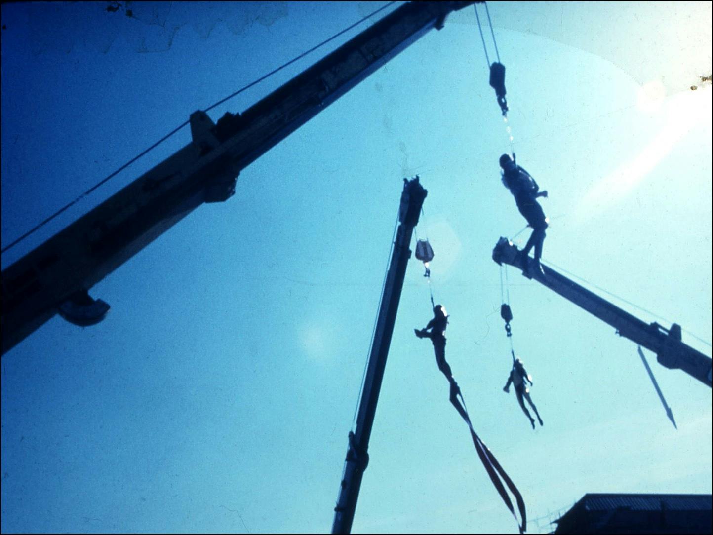 A blue-tinted, high-contrast photograph taken from below. The image shows the silhouettes of three performers, suspended in the air by harnesses attached to the arms of three mobile cranes. The performers hang from their respective cranes at a distance and separated from one another.